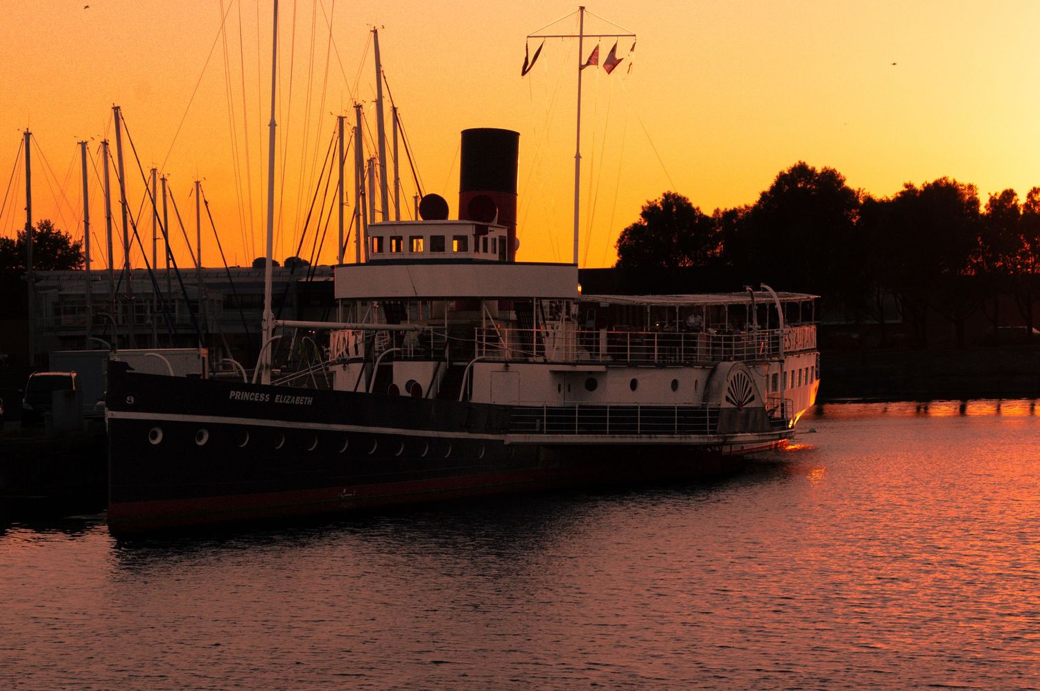 A vintage steamboat docked at a marina during a stunning golden sunset with silhouetted trees in the background.