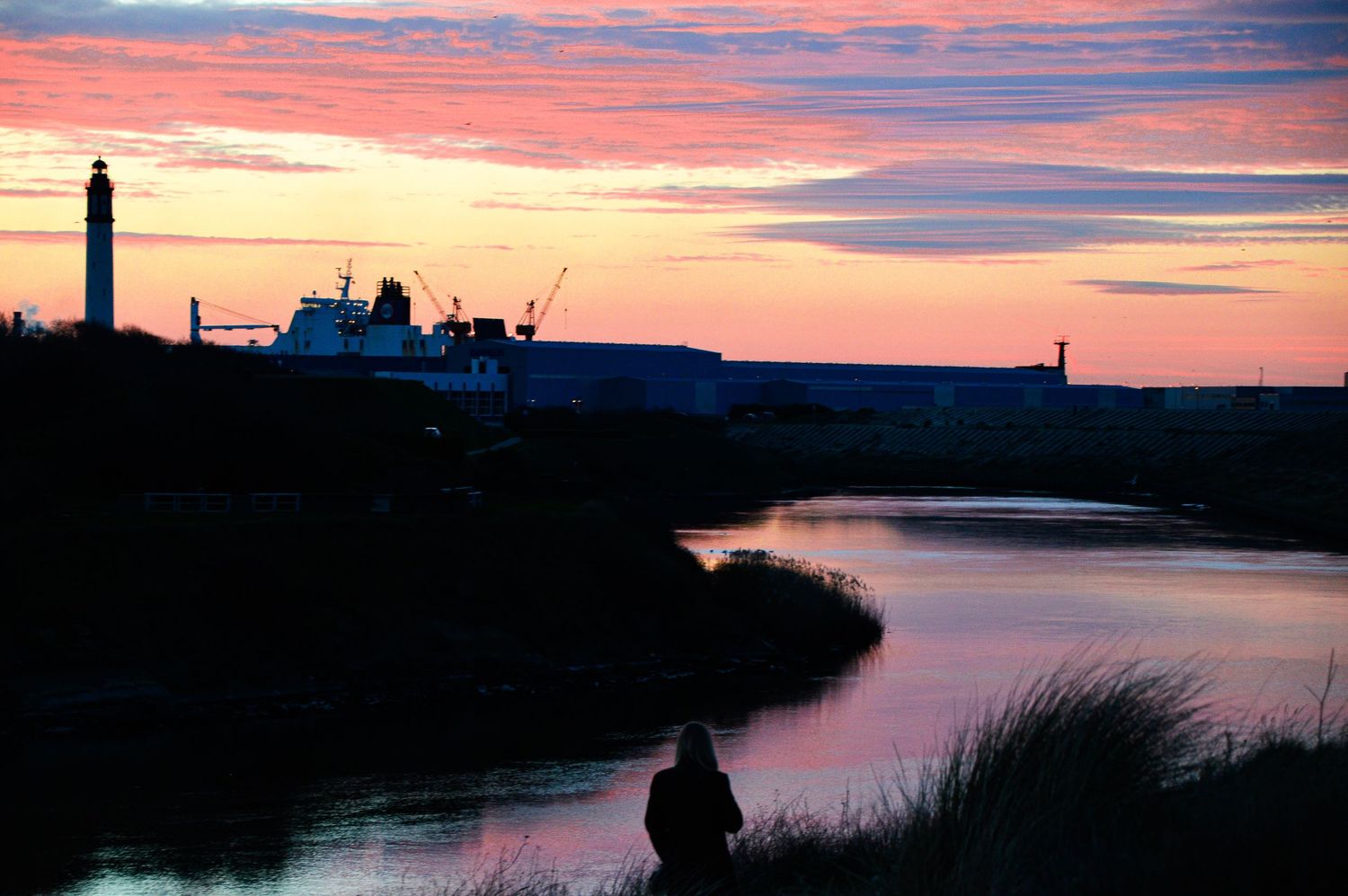 Coastal harbor silhouetted against colorful sunset sky with water reflections.
