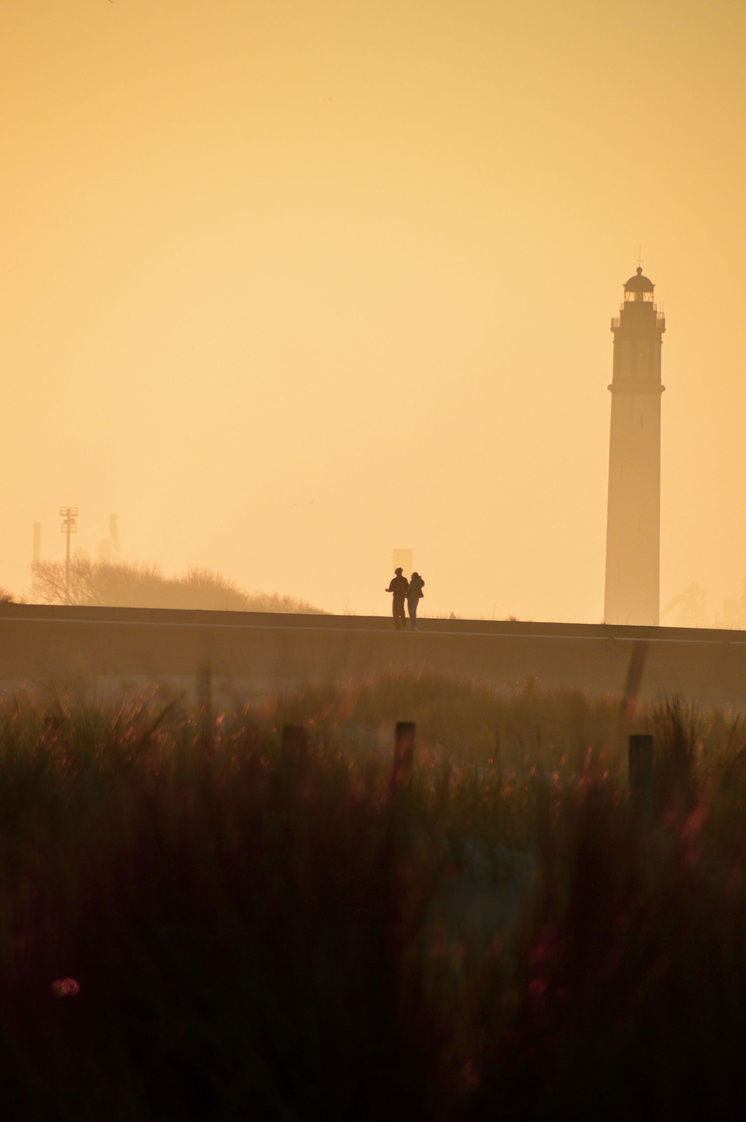 Phare du Risban à Dunkerque et deux silhouettes se détachant sur un ciel doré de coucher de soleil, avec des herbes de plage au premier plan