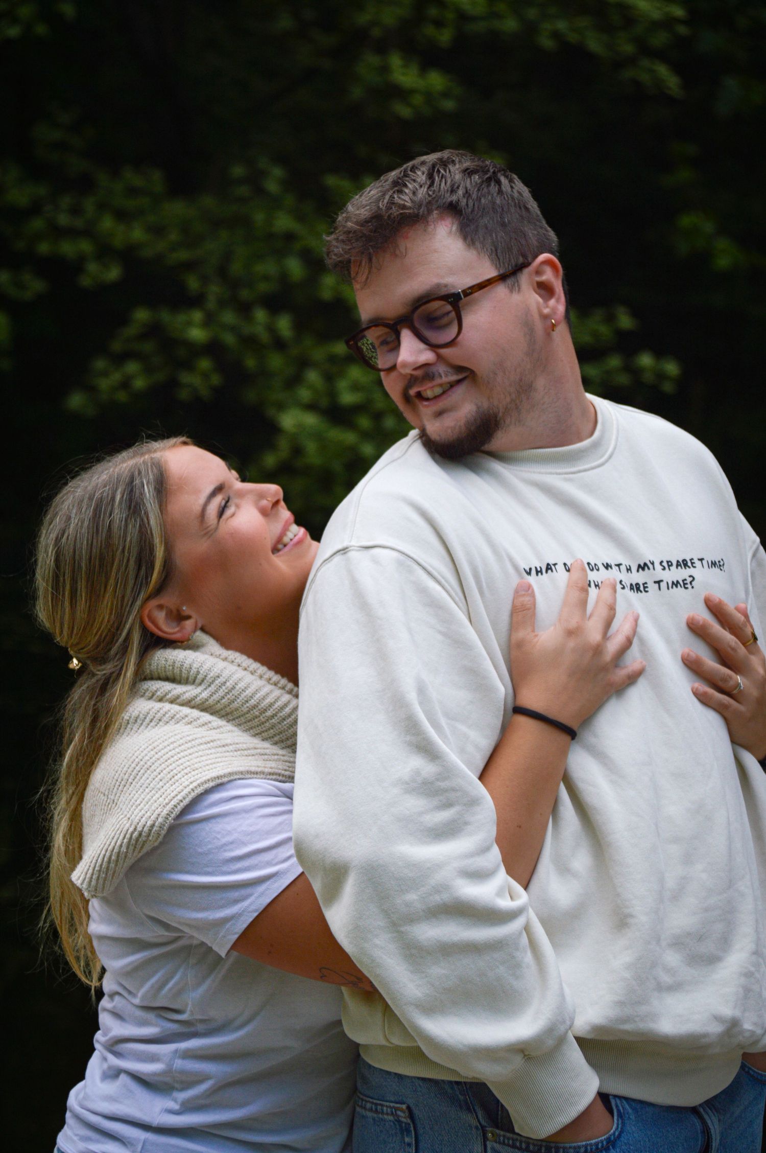 Playful couple embracing and laughing together in white sweaters against natural backdrop.