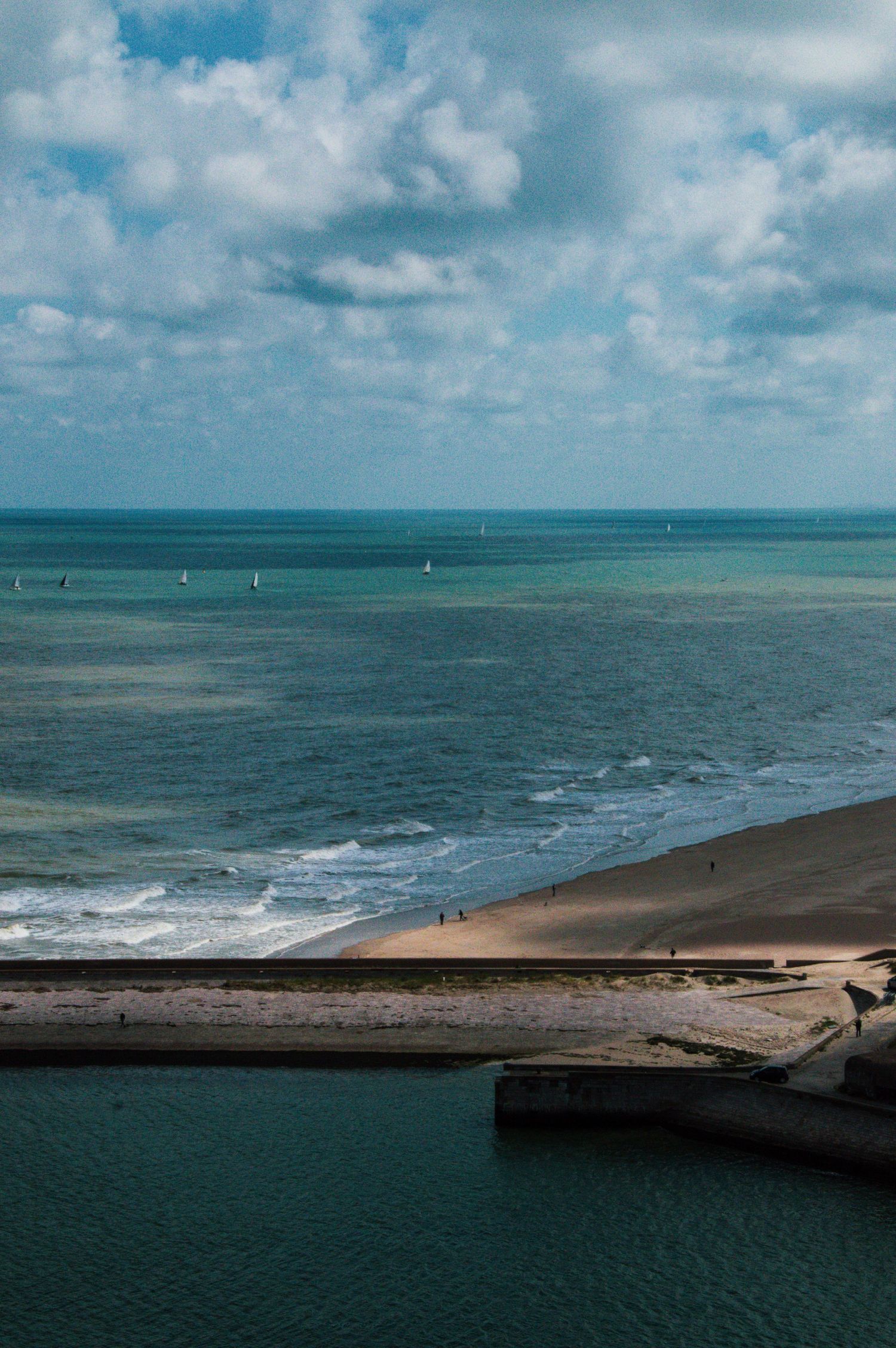 Aerial view of a sandy beach with gentle waves, sailboats on the horizon, and a cloudy blue sky over calm ocean waters.