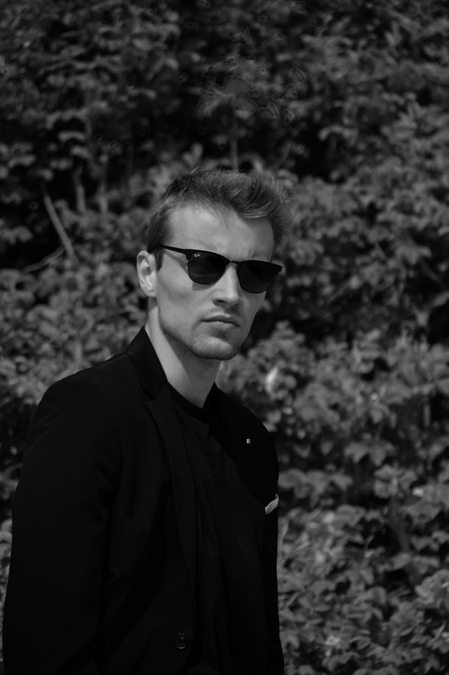 Person in sunglasses and dark clothing photographed in black and white against foliage.
