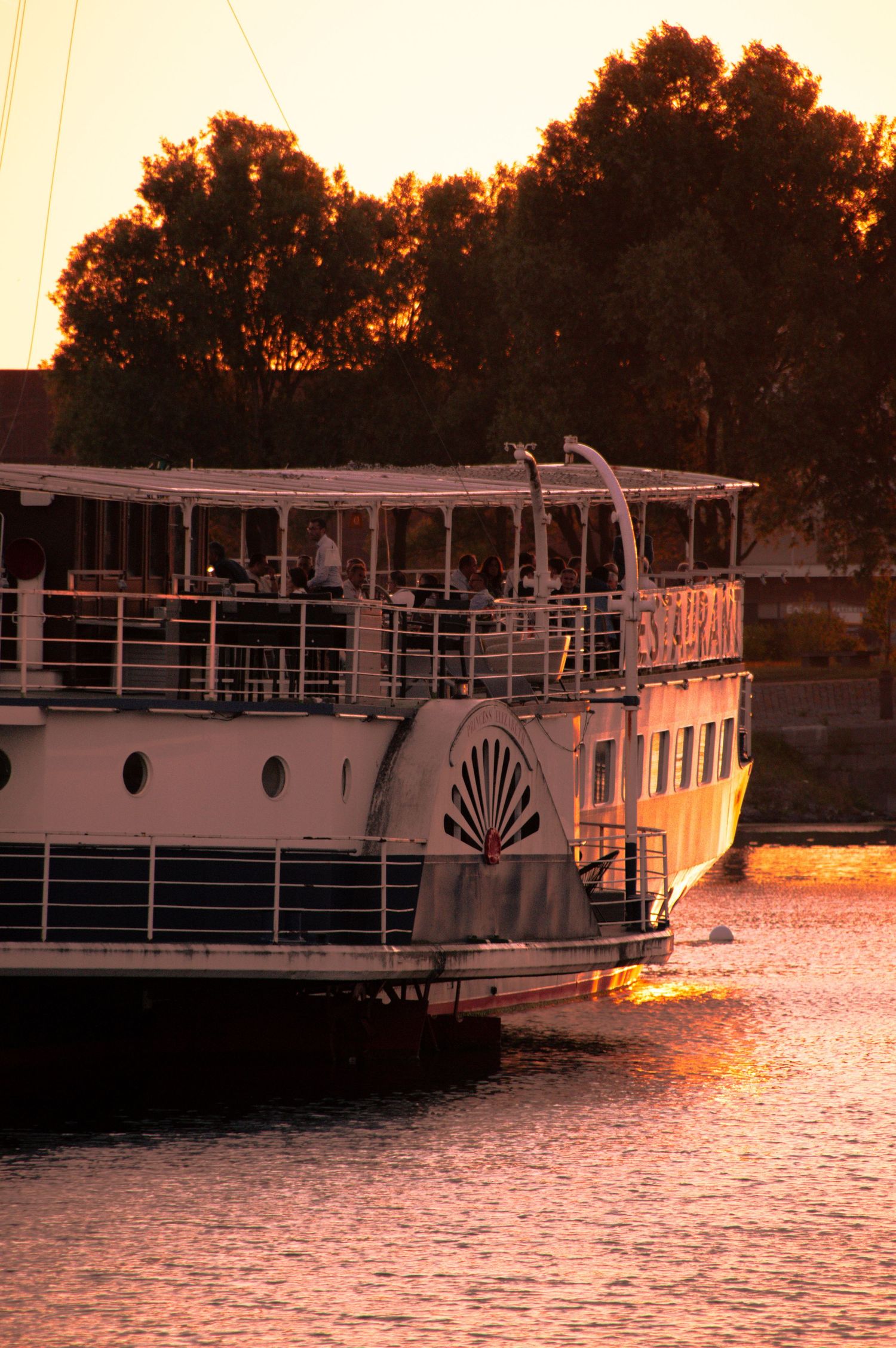 A white paddle steamer cruise boat sailing at sunset with passengers on deck, golden light reflecting on calm water.