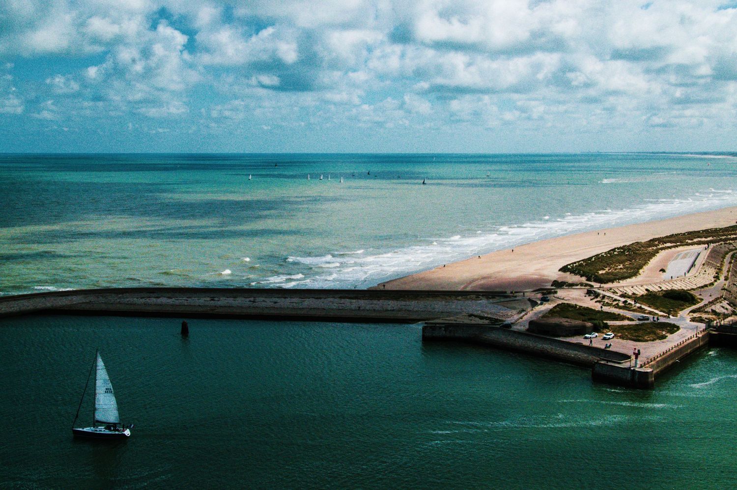 Aerial view of a sandy beach coastline with a sailboat on turquoise water near a harbor wall under cloudy skies.