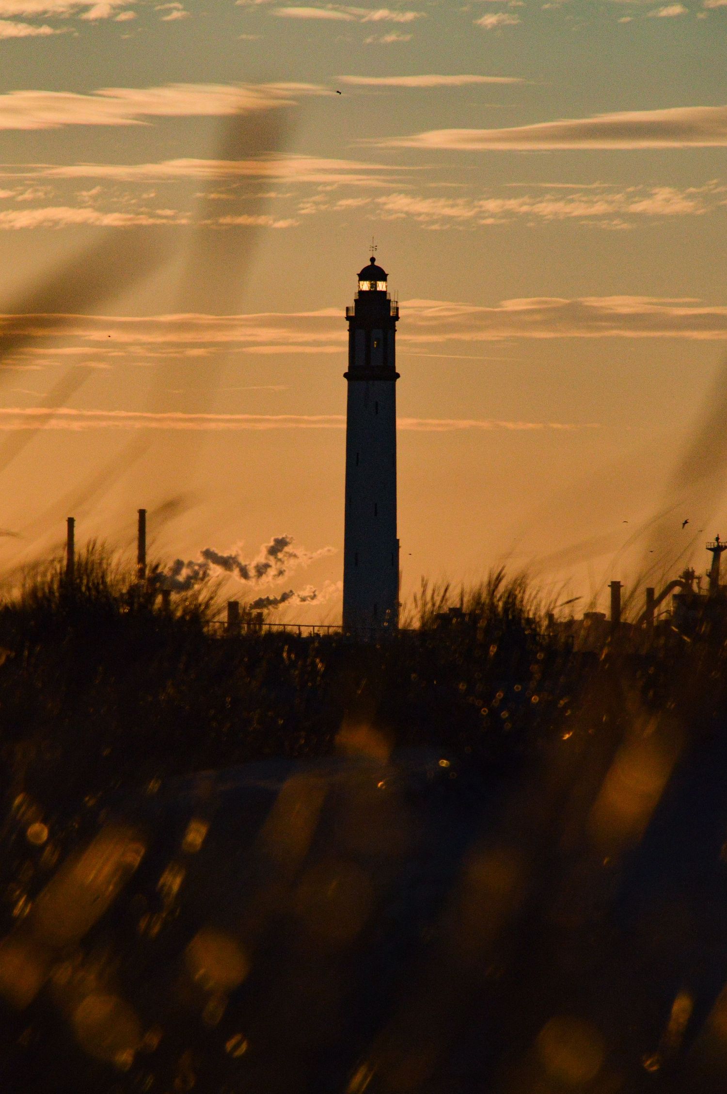 Lighthouse silhouette against warm orange sunset sky with grass in foreground.