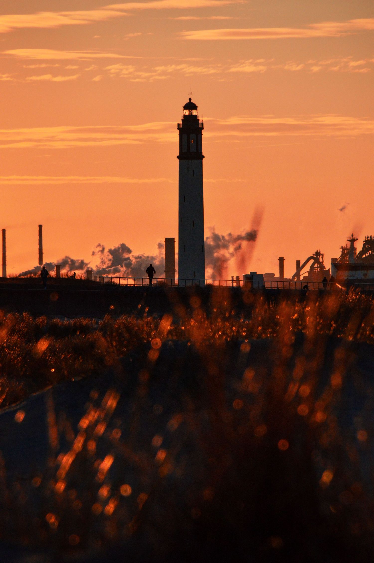 A lighthouse silhouette stands against a vibrant orange sunset sky with industrial structures on the horizon.