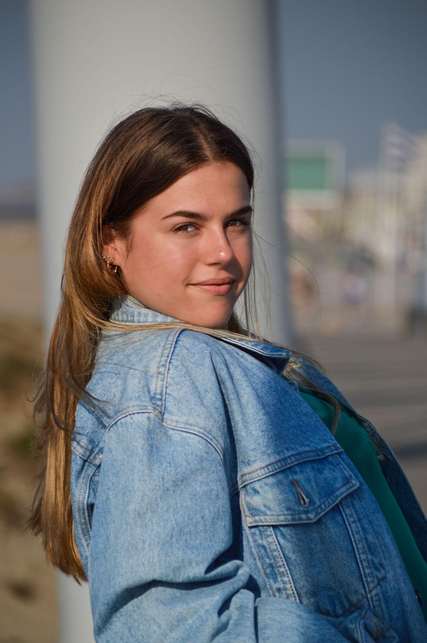 Young woman in denim jacket smiling over her shoulder in warm sunlight.