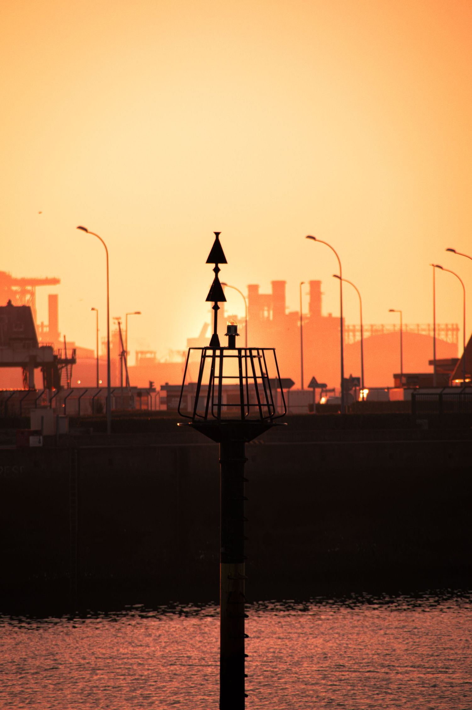 Silhouette of harbor navigation markers and city skyline against vibrant orange sunset sky reflecting on calm waters.