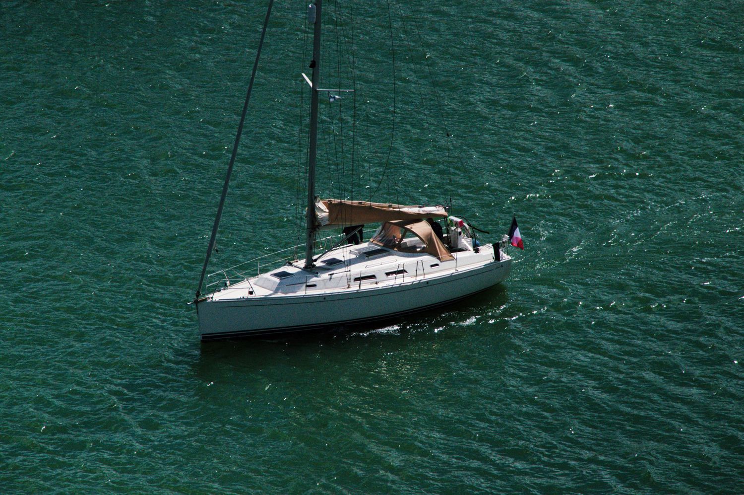 A white sailboat with a mast glides across calm green water, with people relaxing on deck on a sunny day.