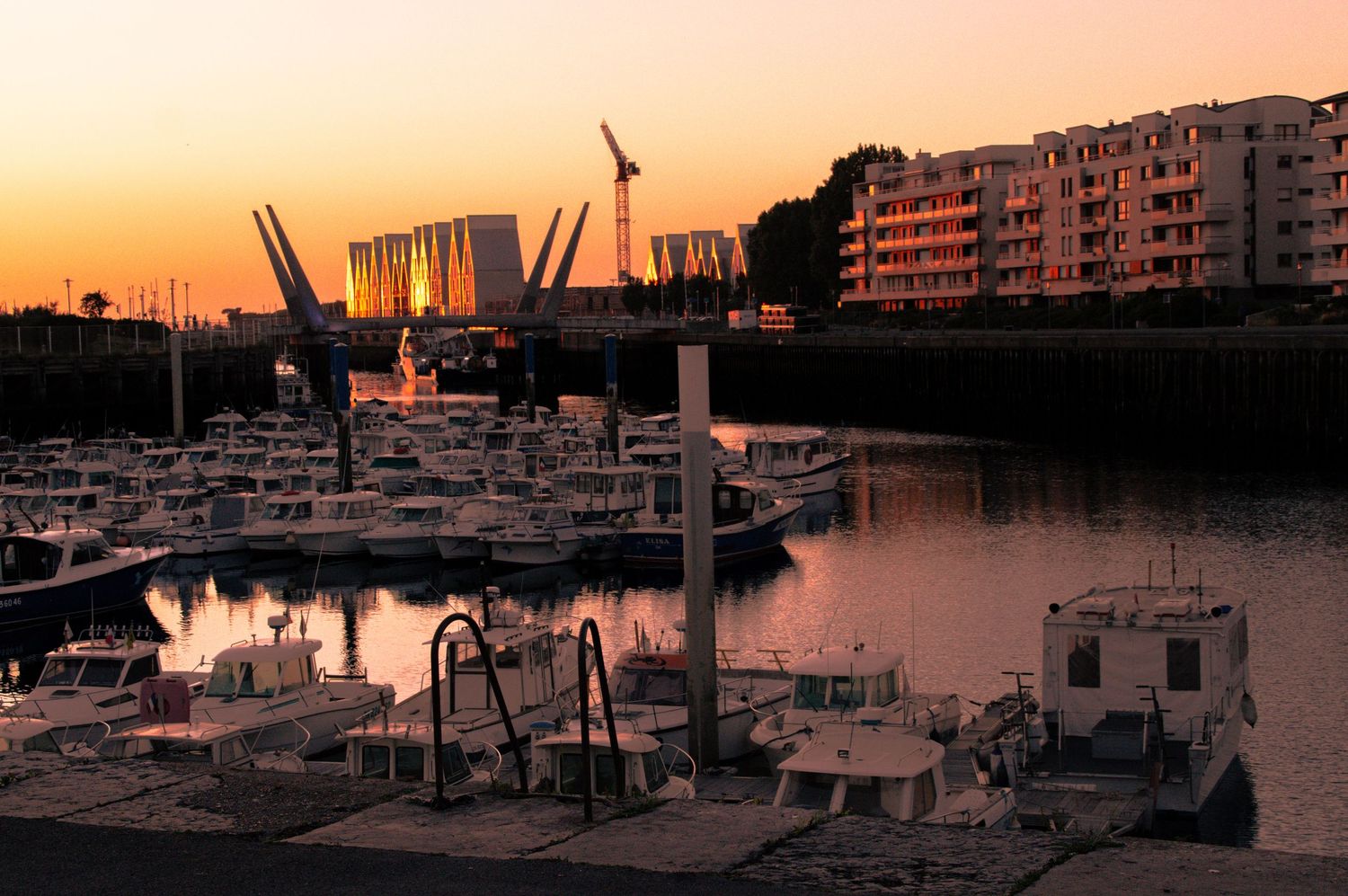Sunset over a marina with boats docked, cranes silhouetted against orange sky, and waterfront apartments reflected in calm water.