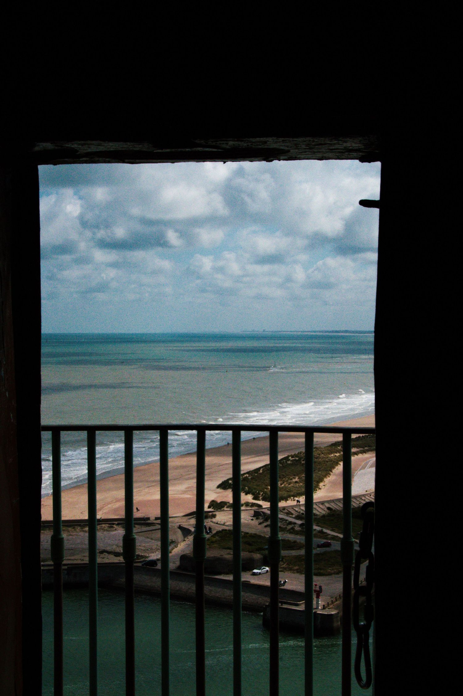 View through a dark doorway with metal railings overlooking a sandy beach and cloudy sky over the ocean.
