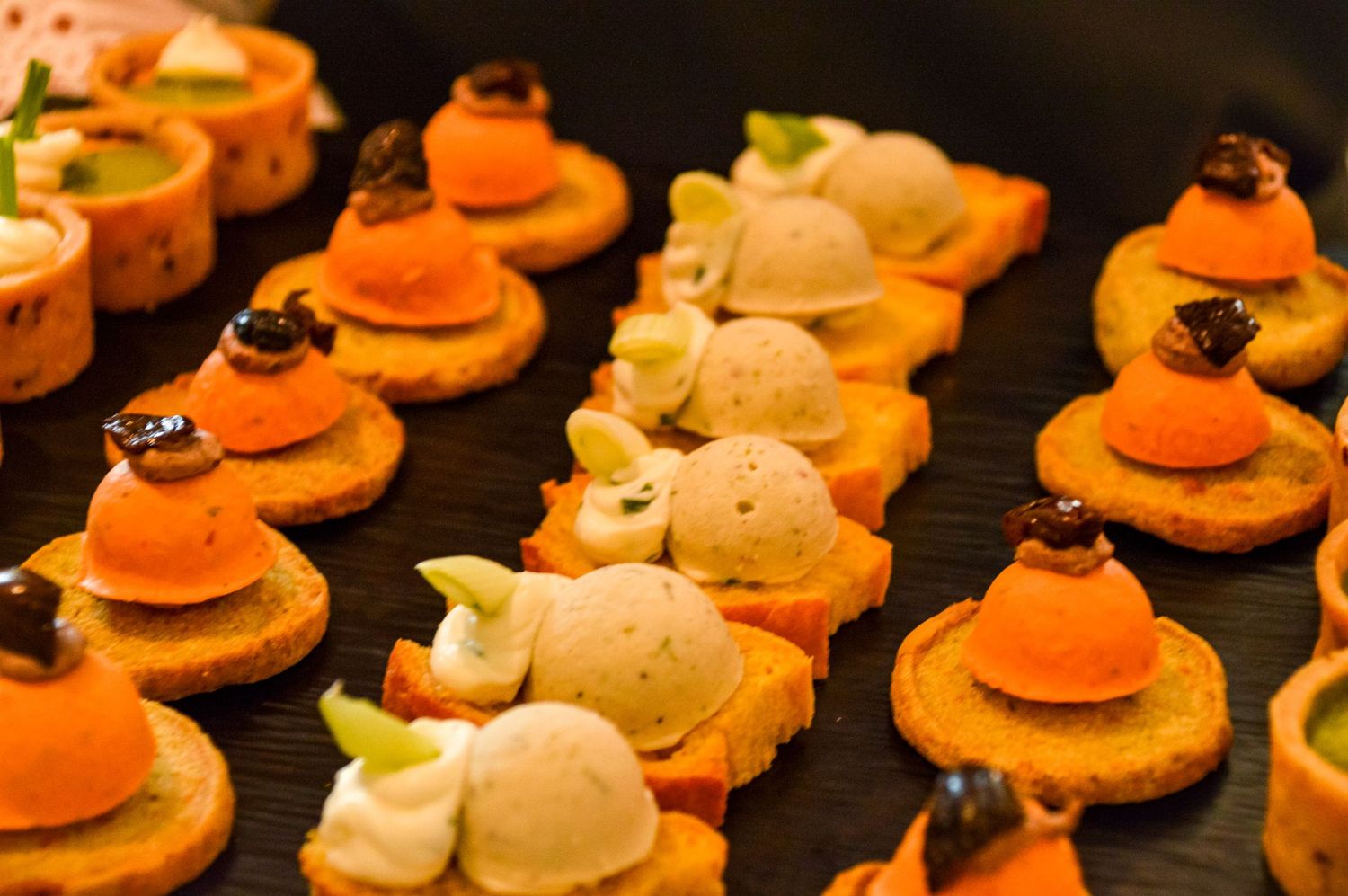 Elegant canapés with orange and white toppings arranged in neat rows on dark platter.