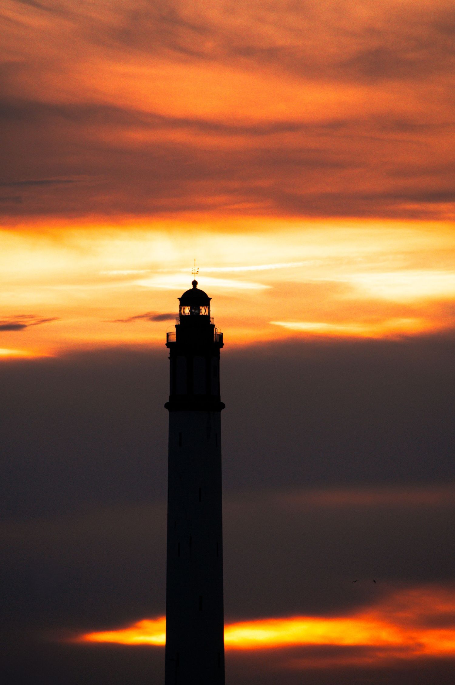 Silhouette of a tall lighthouse against a dramatic orange and red sunset sky with glowing clouds.