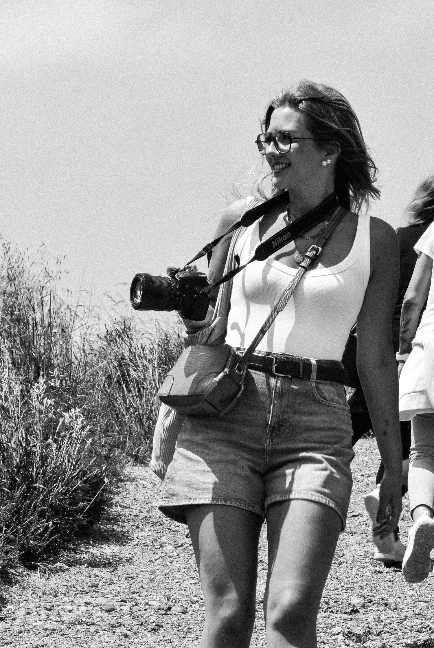 Black and white photo of people walking on a trail wearing casual summer clothing and carrying bags.