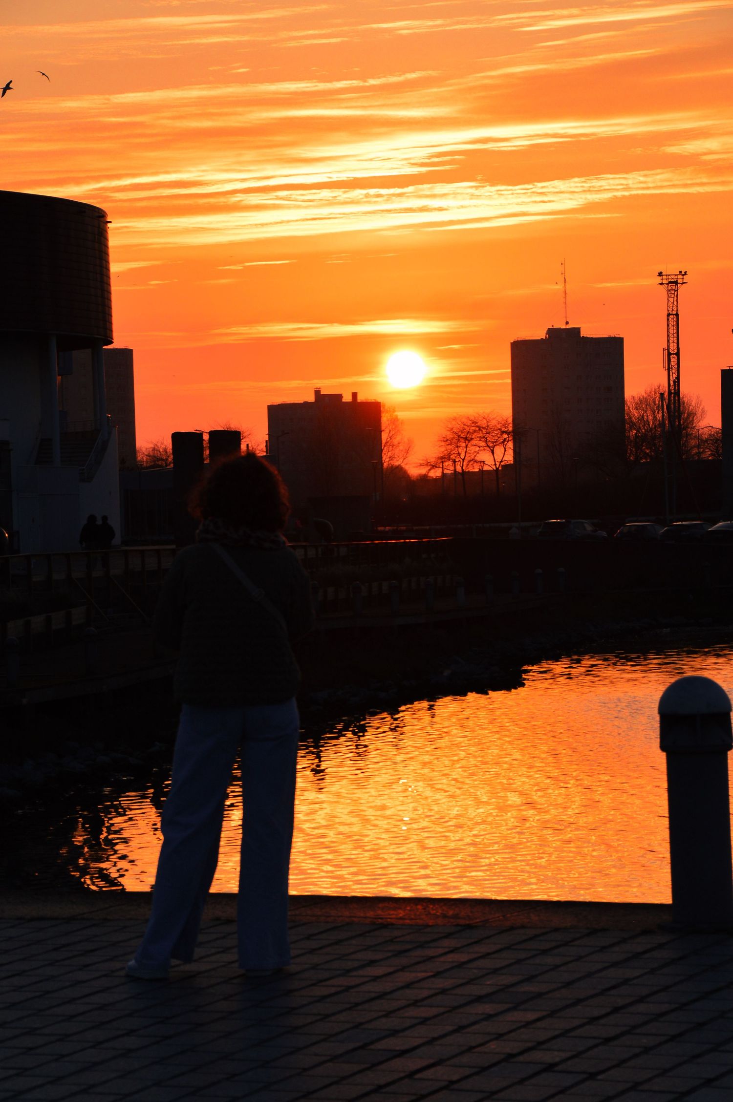 Front de mer urbain en silhouette sur un coucher de soleil orange intense, avec des bâtiments et leurs reflets dans l’eau.