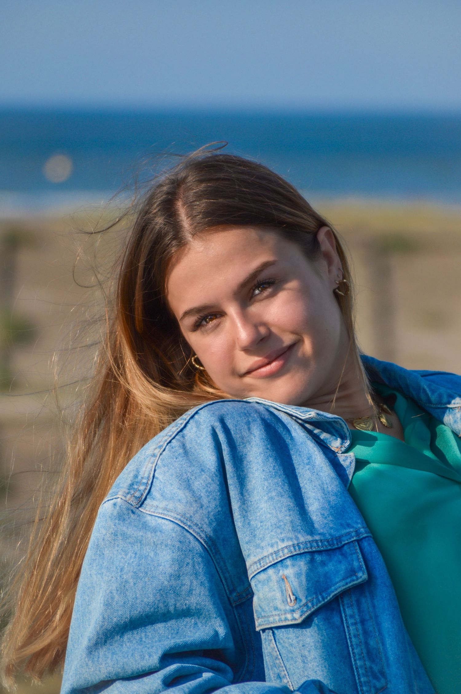 Woman in denim jacket and green top poses against blue ocean backdrop.