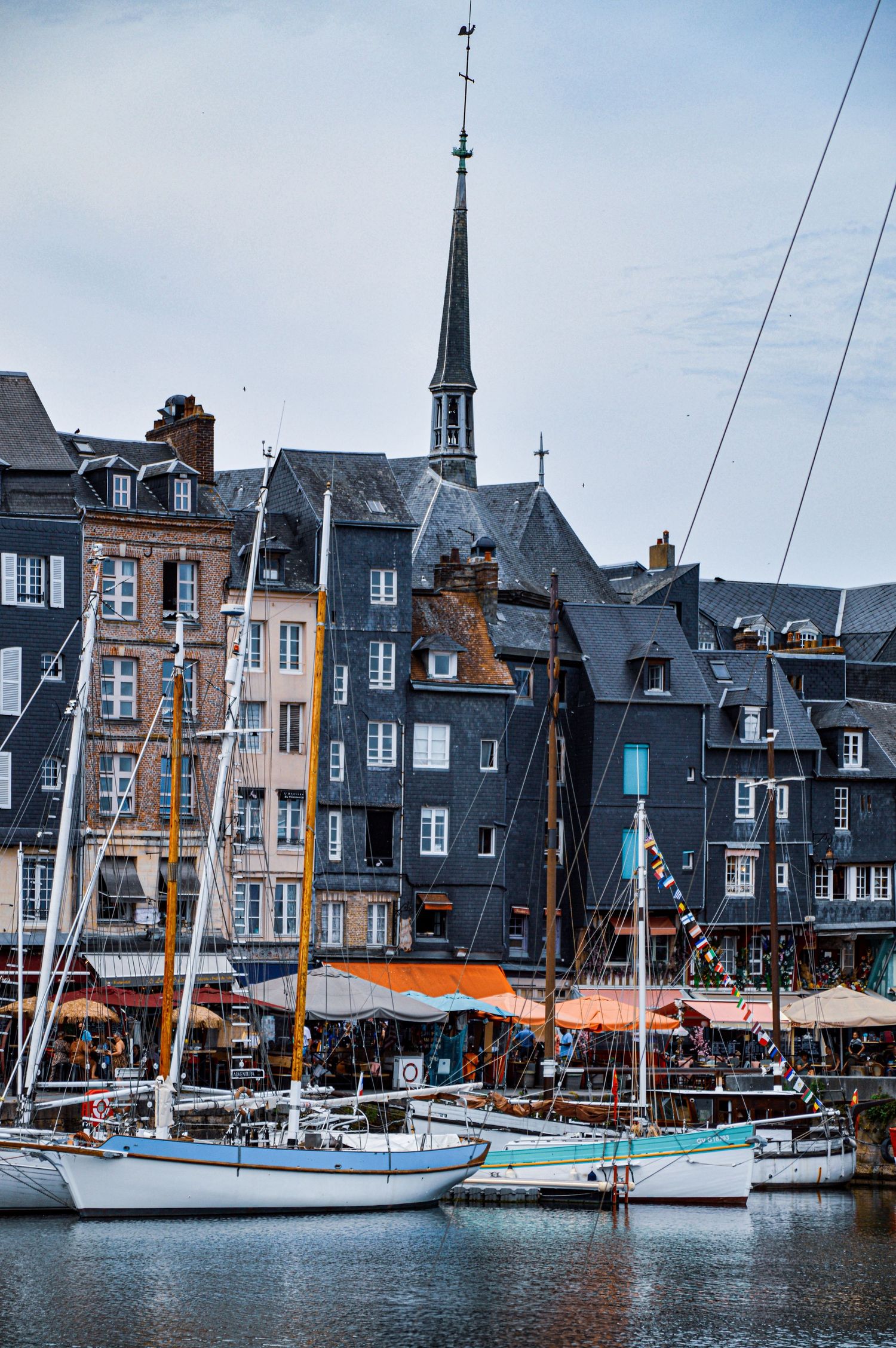 Historic harbor with sailboats moored along colorful buildings and church spires in Honfleur, France.