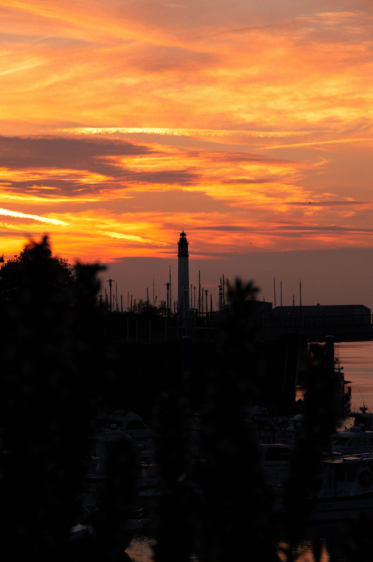 Stunning orange and red sunset silhouettes a lighthouse and marina with boats against a dramatic cloudy sky.