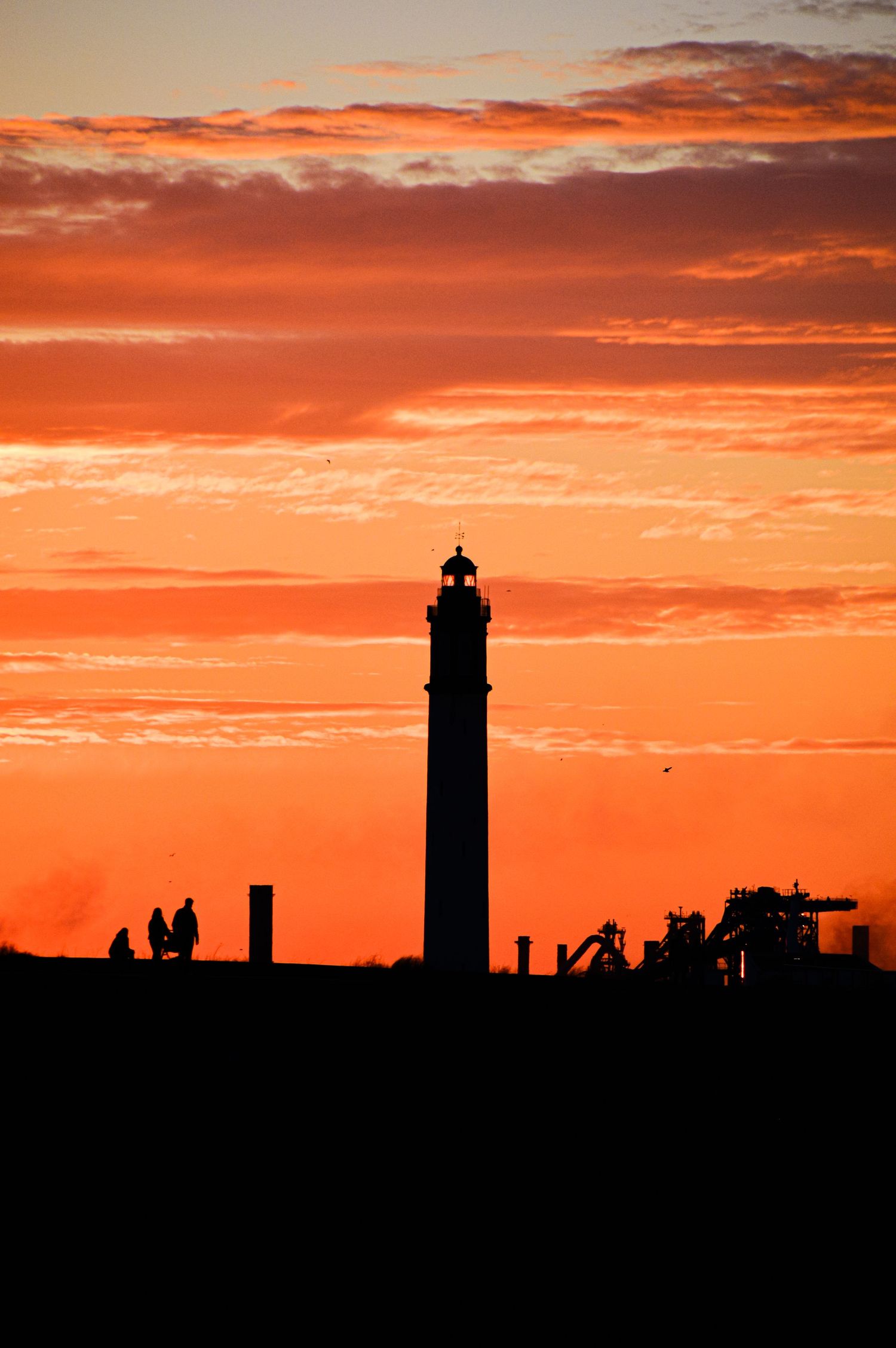 Industrial skyline with lighthouse silhouetted against dramatic orange sunset.