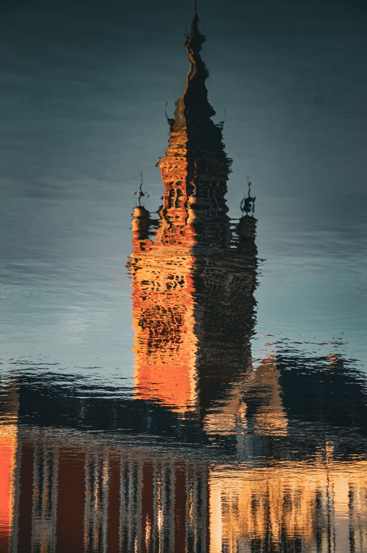 Orange-tinted clock tower reflection rippling in dark harbor waters at sunset.