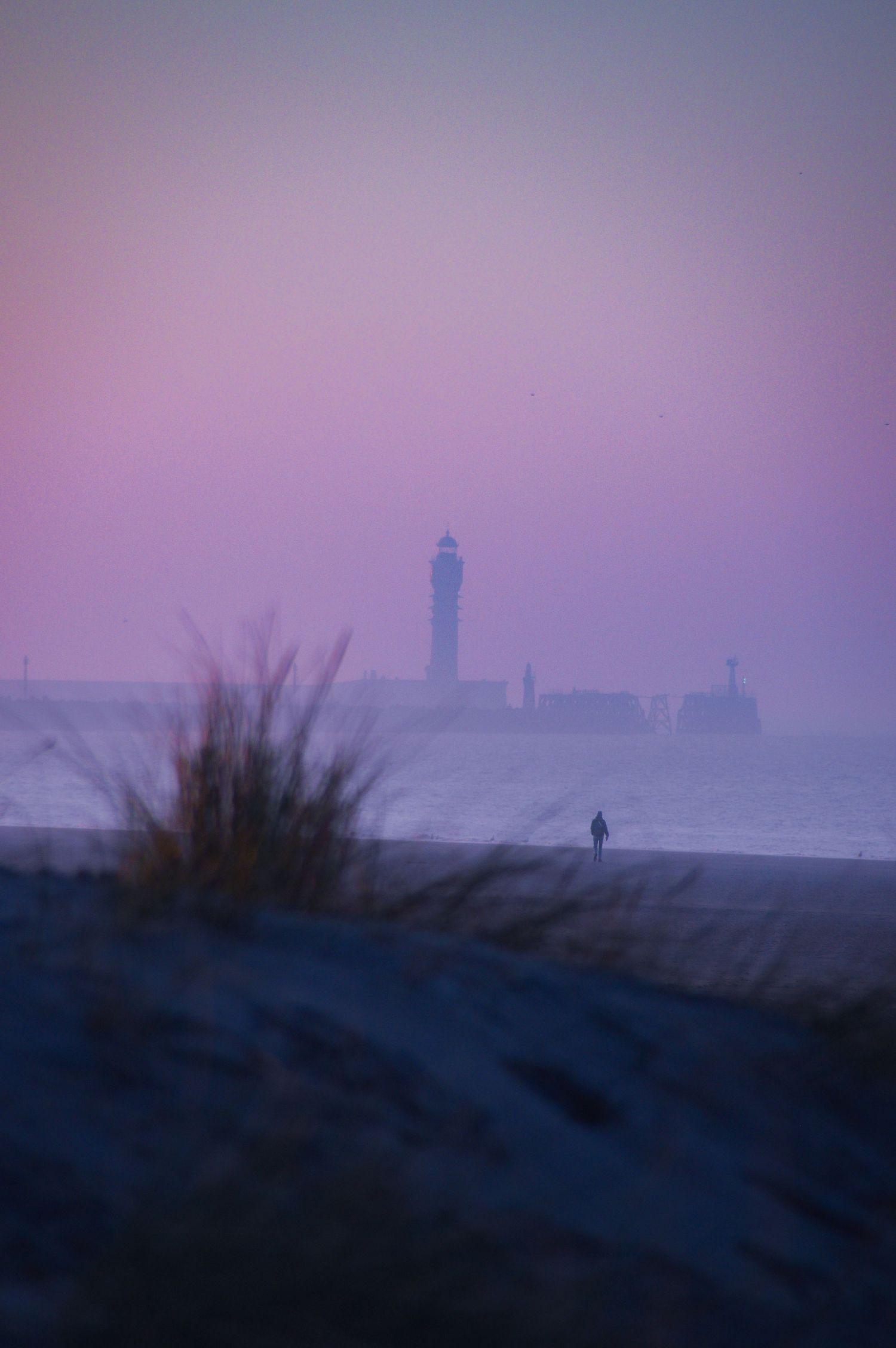 Phare brumeux sur la côte au crépuscule, avec une silhouette marchant le long de la plage dans une lumière violette du soir.
