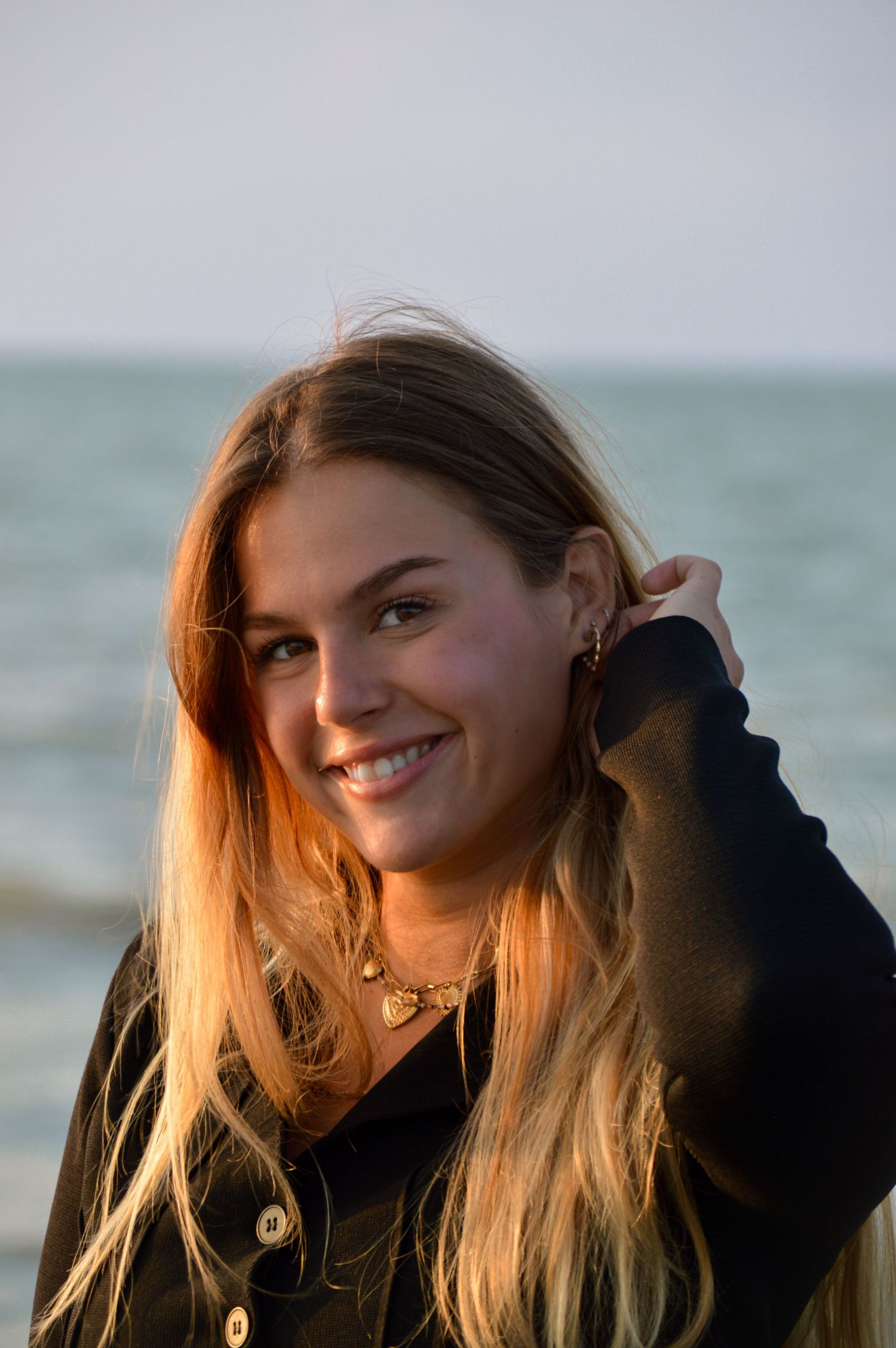 Person in black hoodie smiling by the ocean during golden hour.