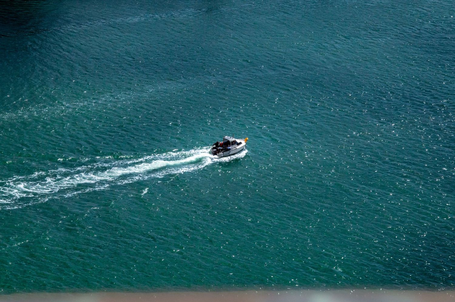 Aerial view of a small motorboat cruising through turquoise blue-green water, leaving a white wake trail behind.