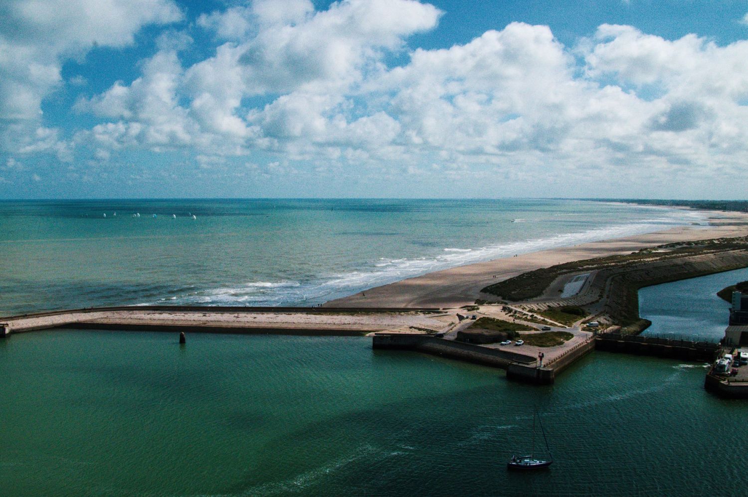 Aerial view of a coastal harbor with a long breakwater, sandy beach, and turquoise ocean waters under a cloudy sky.