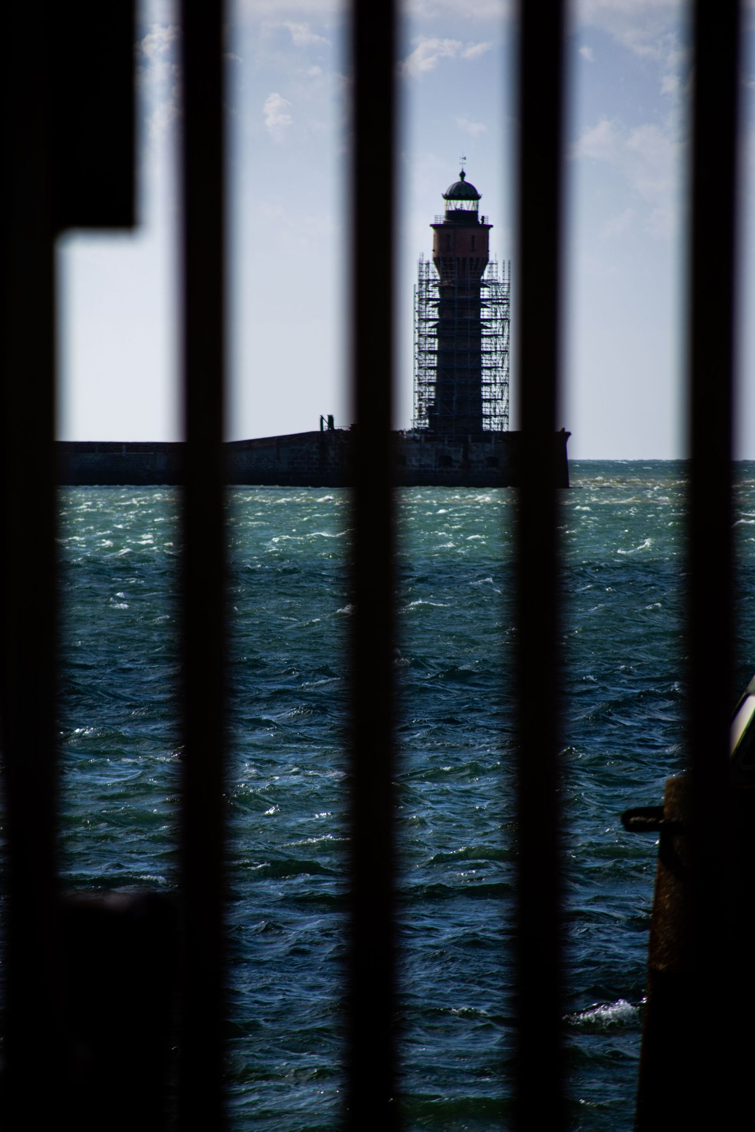 A lighthouse under scaffolding viewed through dark metal bars, with choppy green sea waters under a partly cloudy sky.