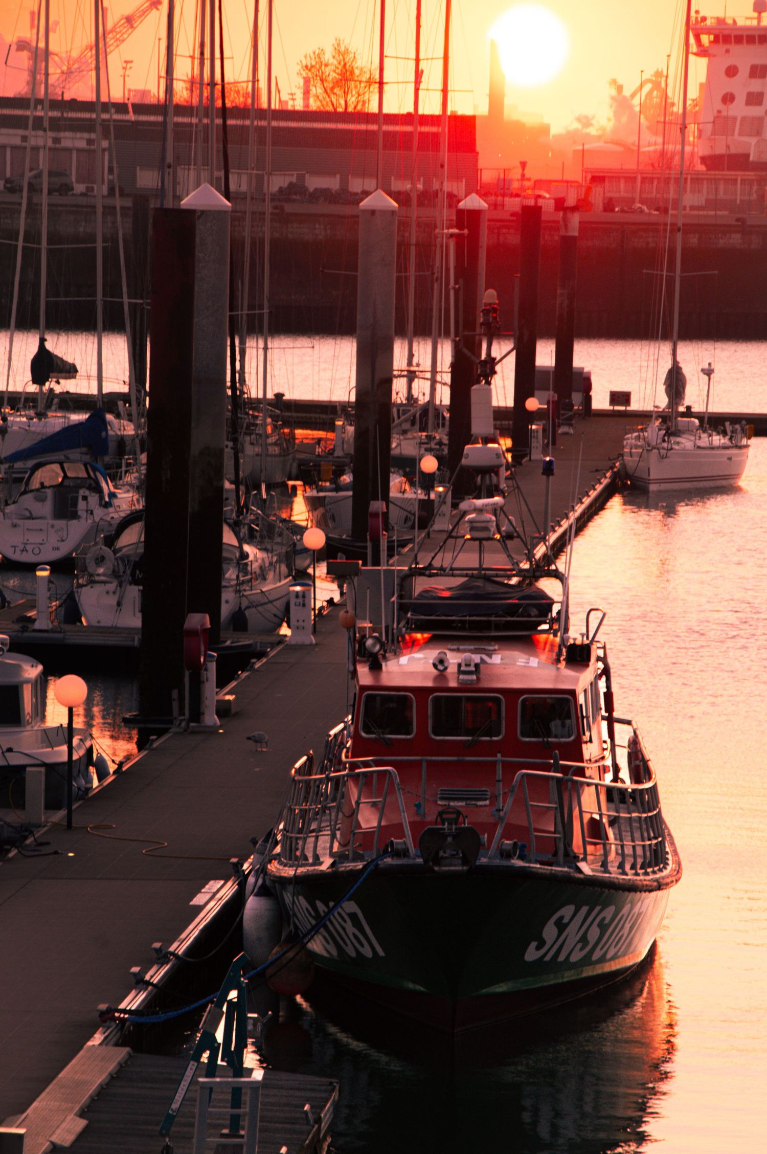 A Coast Guard vessel docked at marina during dramatic orange sunset with sailboats and harbor posts silhouetted against sky.