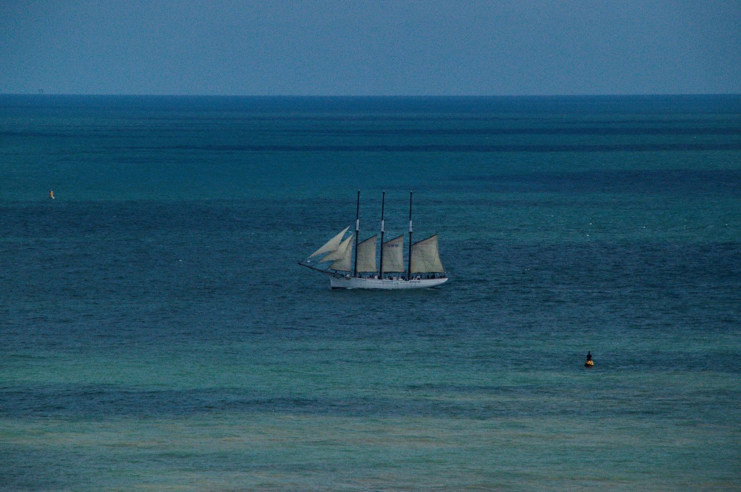 A tall ship with white sails sailing on calm turquoise ocean waters under a clear blue sky.