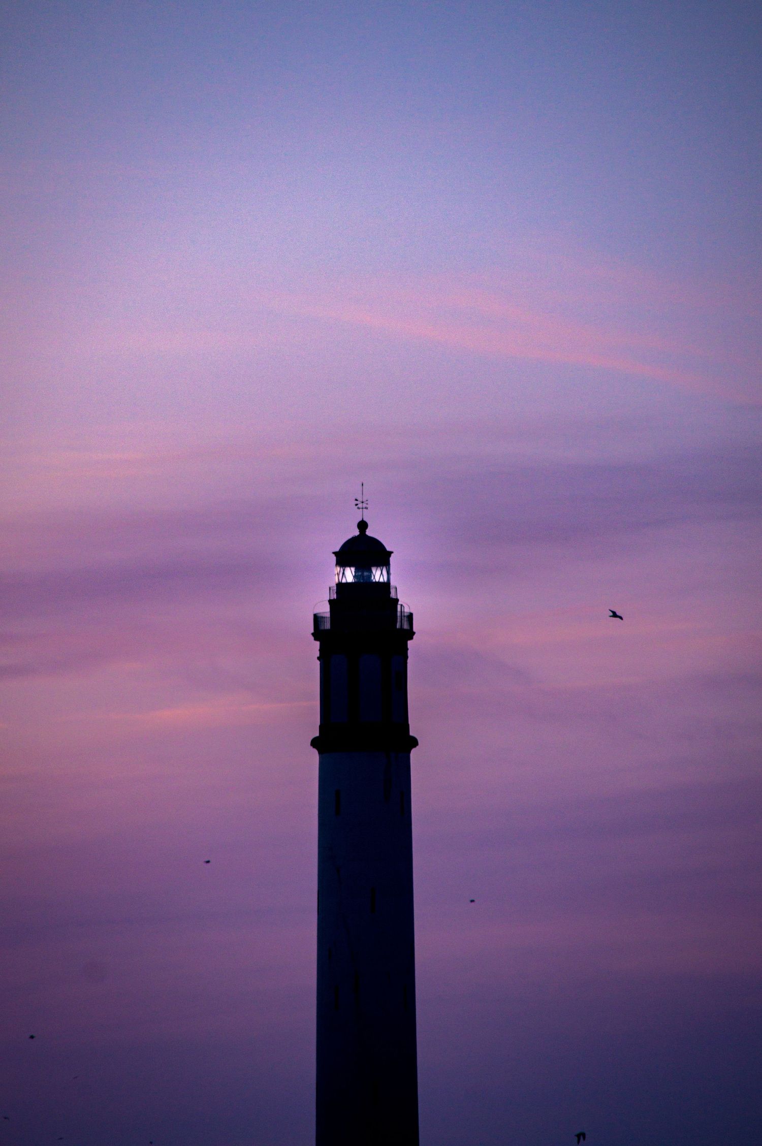 Silhouette of lighthouse tower against vibrant purple and pink sunset sky with birds flying in the distance.