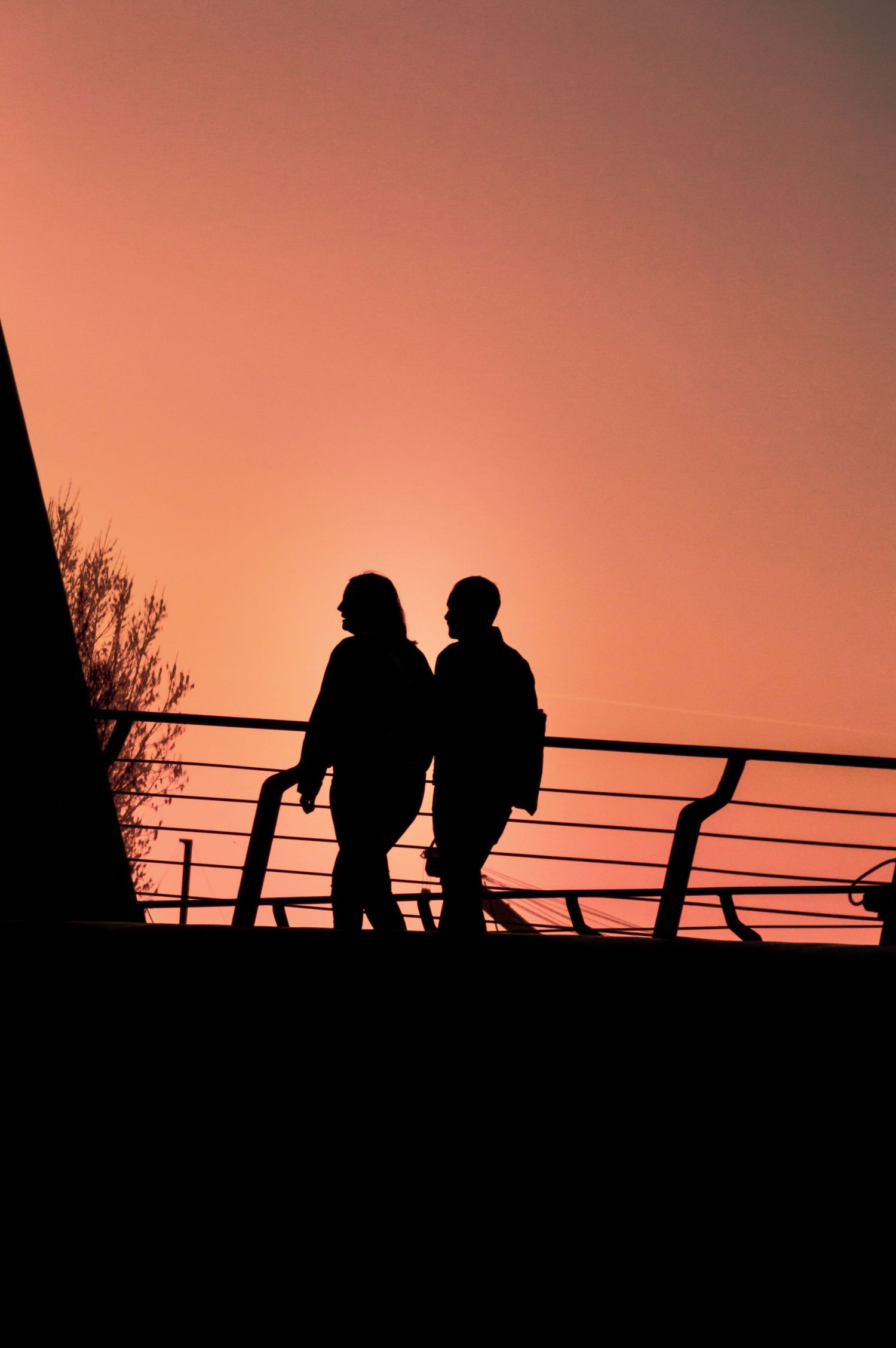Silhouettes of people walking along a railing against a vibrant orange sunset sky.
