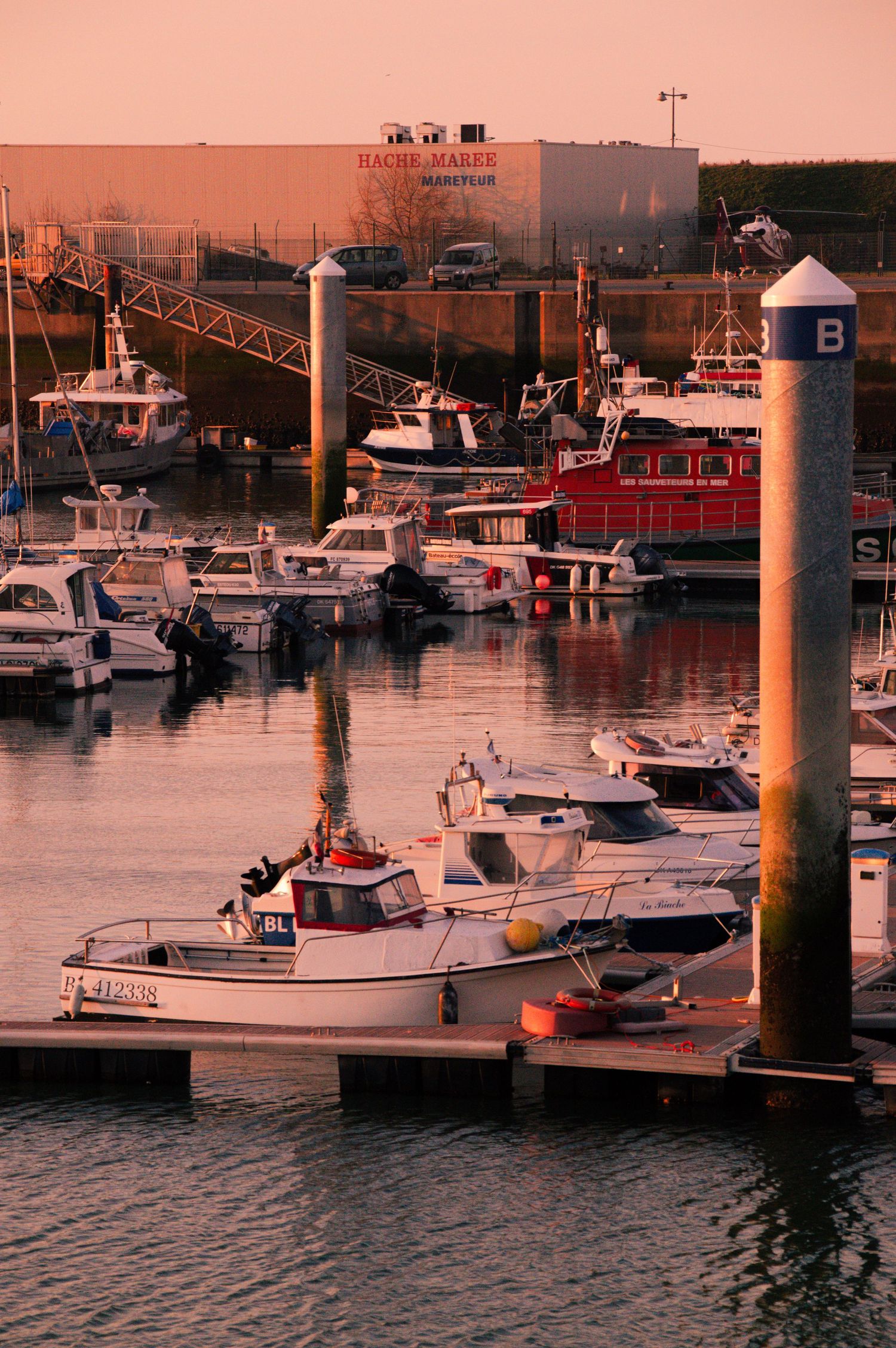 Boats docked at marina during sunset with warm orange glow reflecting off water and industrial buildings in background.