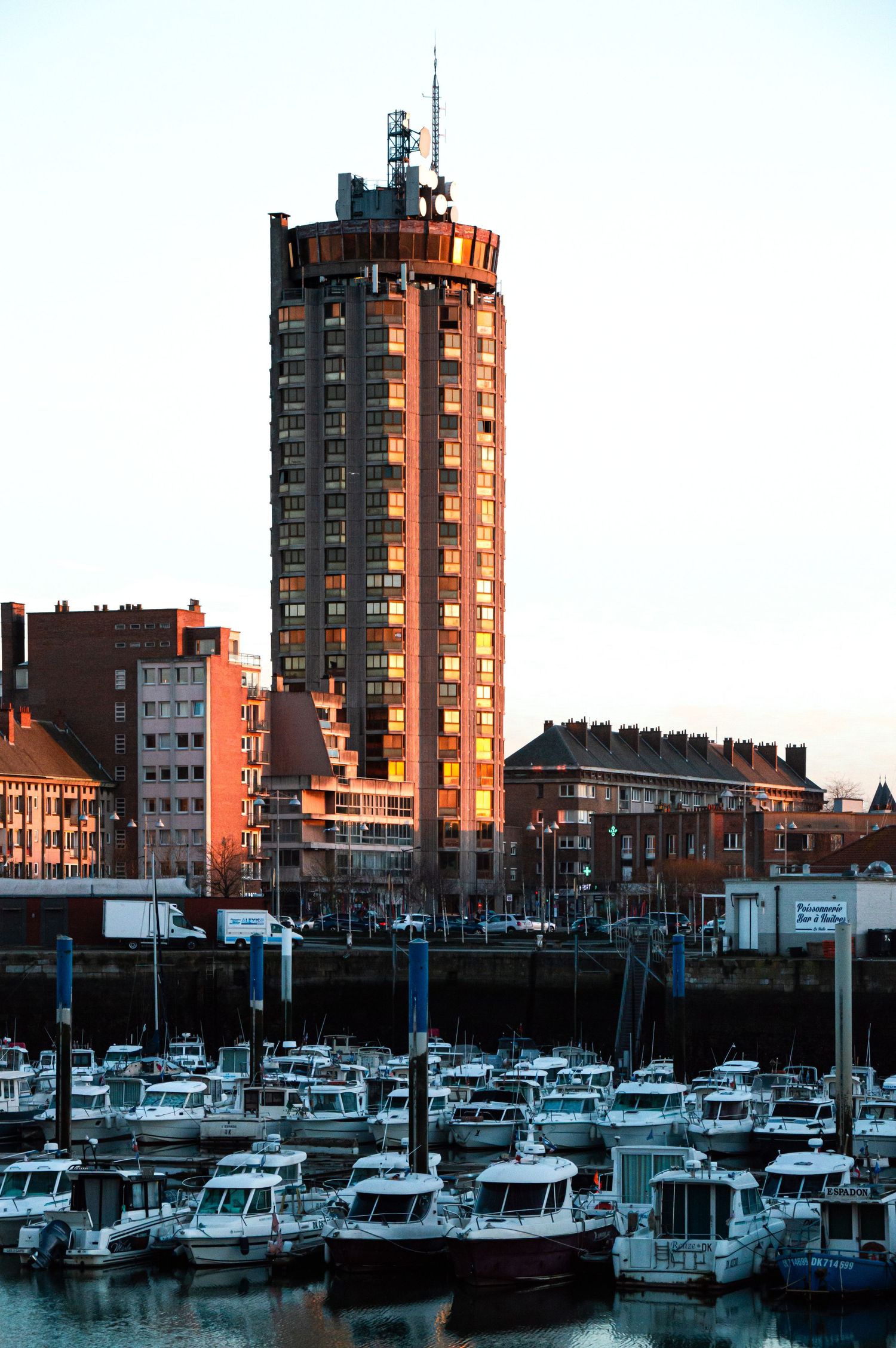 Marina with boats docked alongside tall residential buildings during sunset creating warm orange reflections.