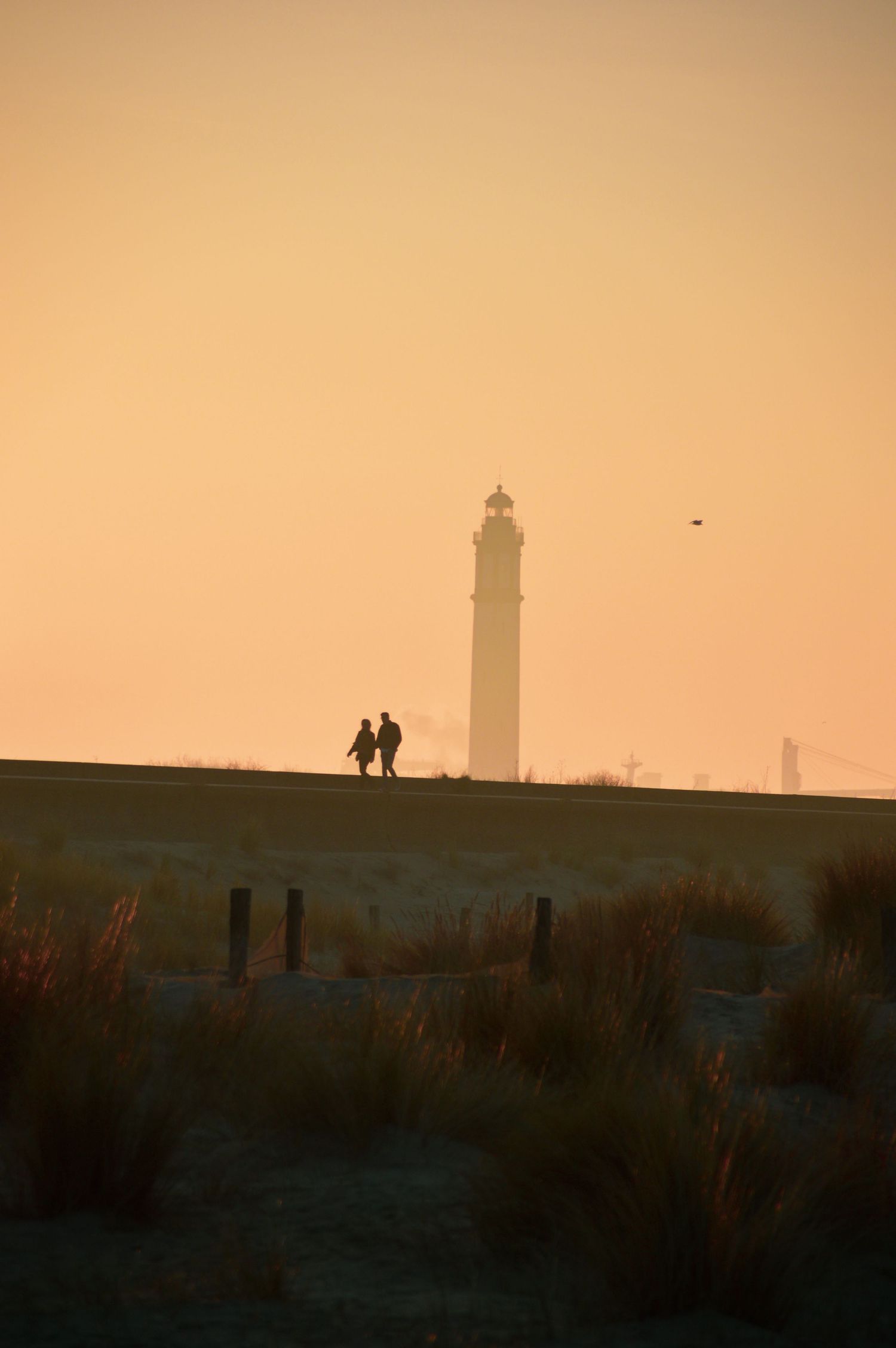 Deux silhouettes se tiennent près du phare du Risban à Dunkerque sur fond de ciel de coucher de soleil orange et chaleureux.