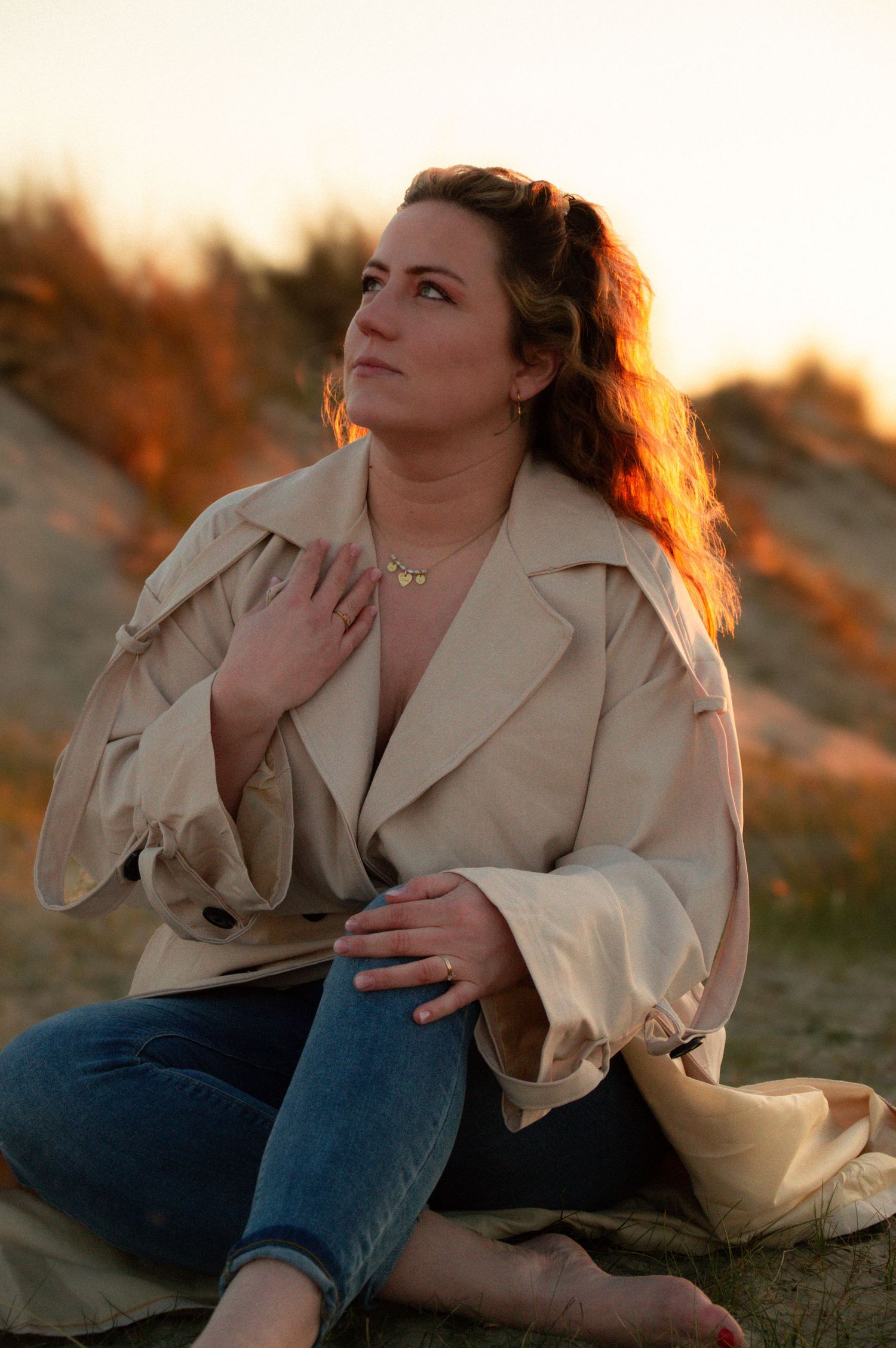 Woman in cream jacket sitting cross-legged on beach sand gazing upward at golden sunset.