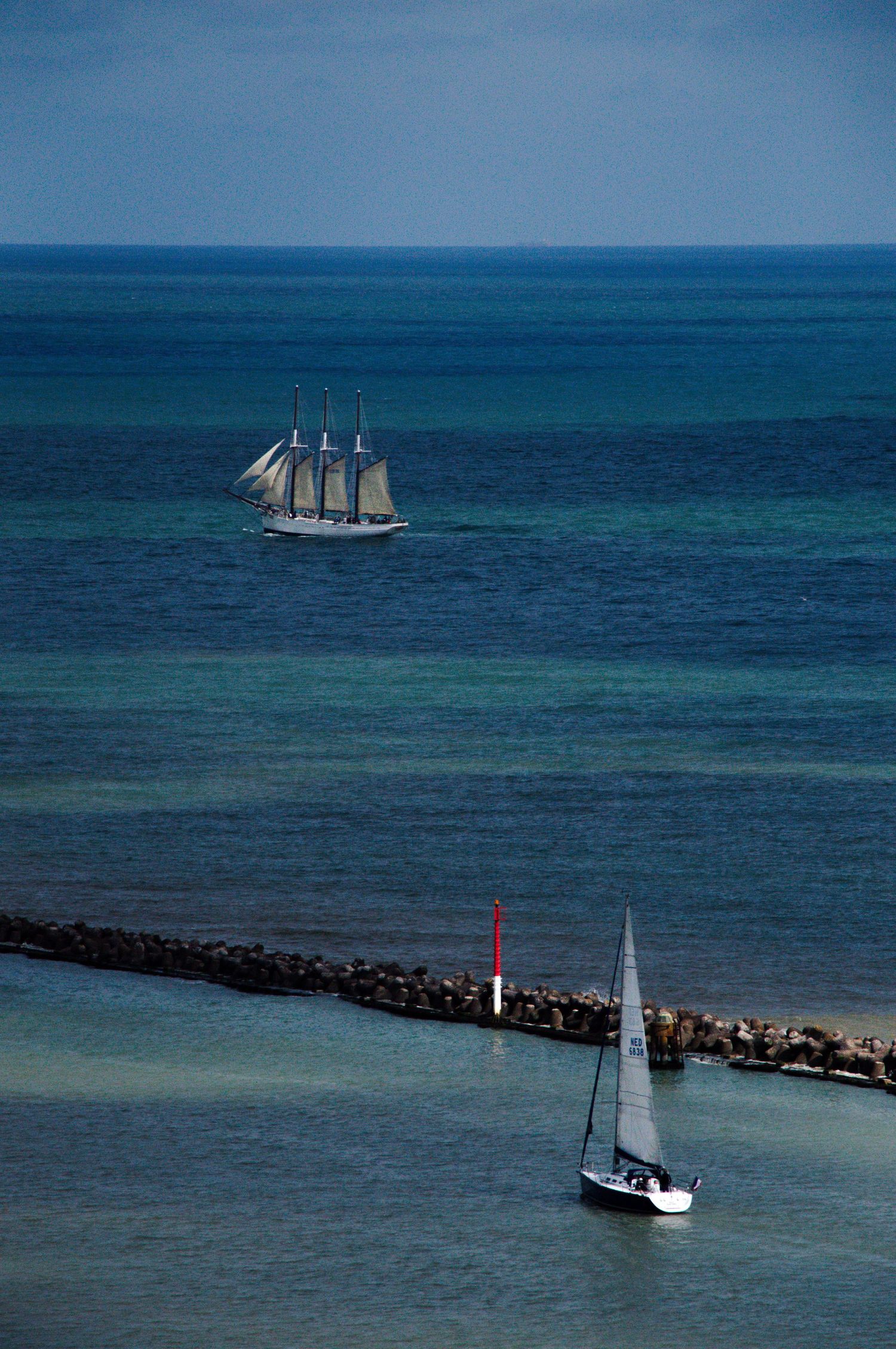 A tall ship with white sails and a small sailboat near a rocky breakwater on calm blue ocean waters.