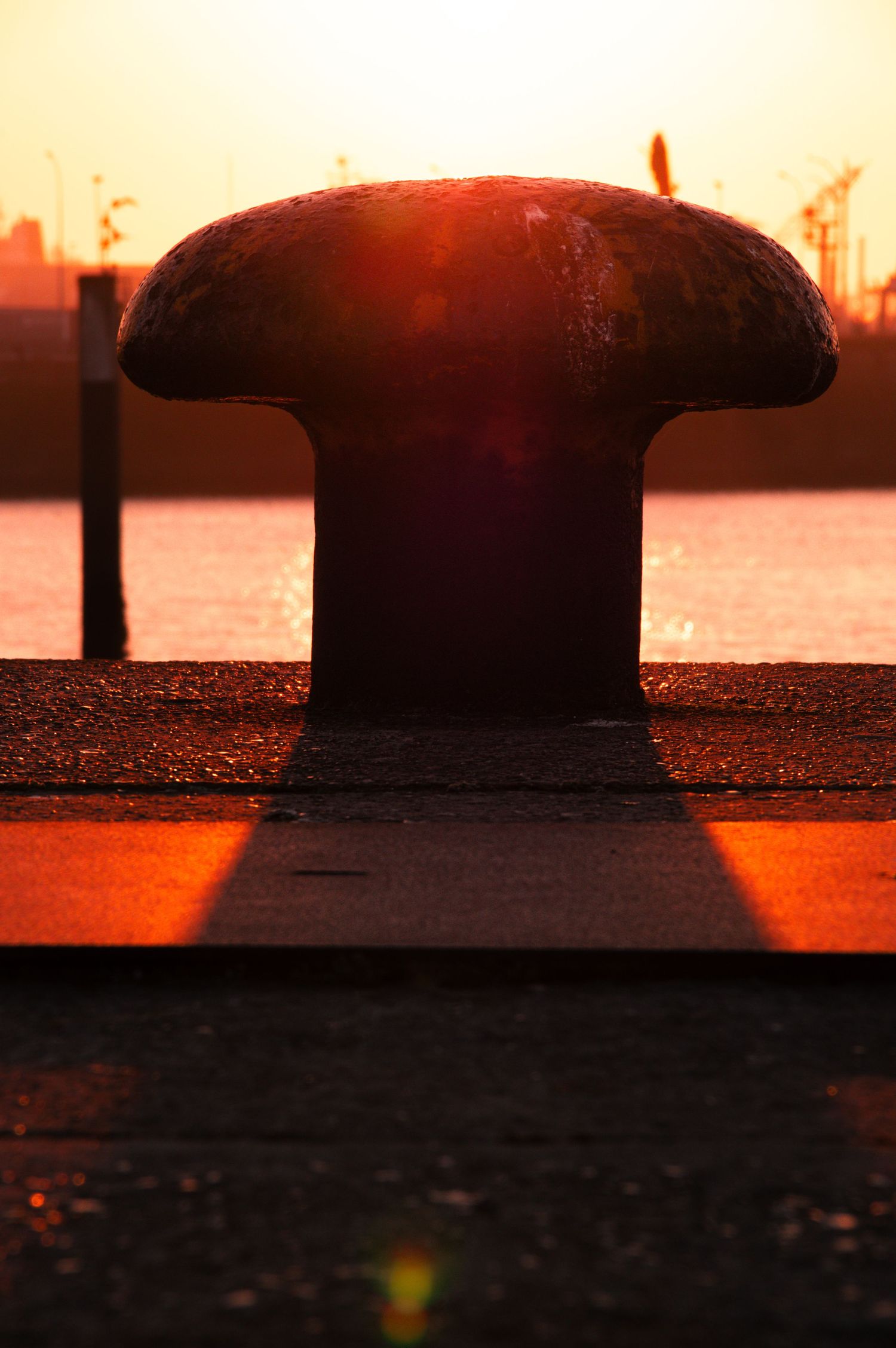 A silhouetted mooring bollard against an orange sunset sky reflecting on calm harbor waters.