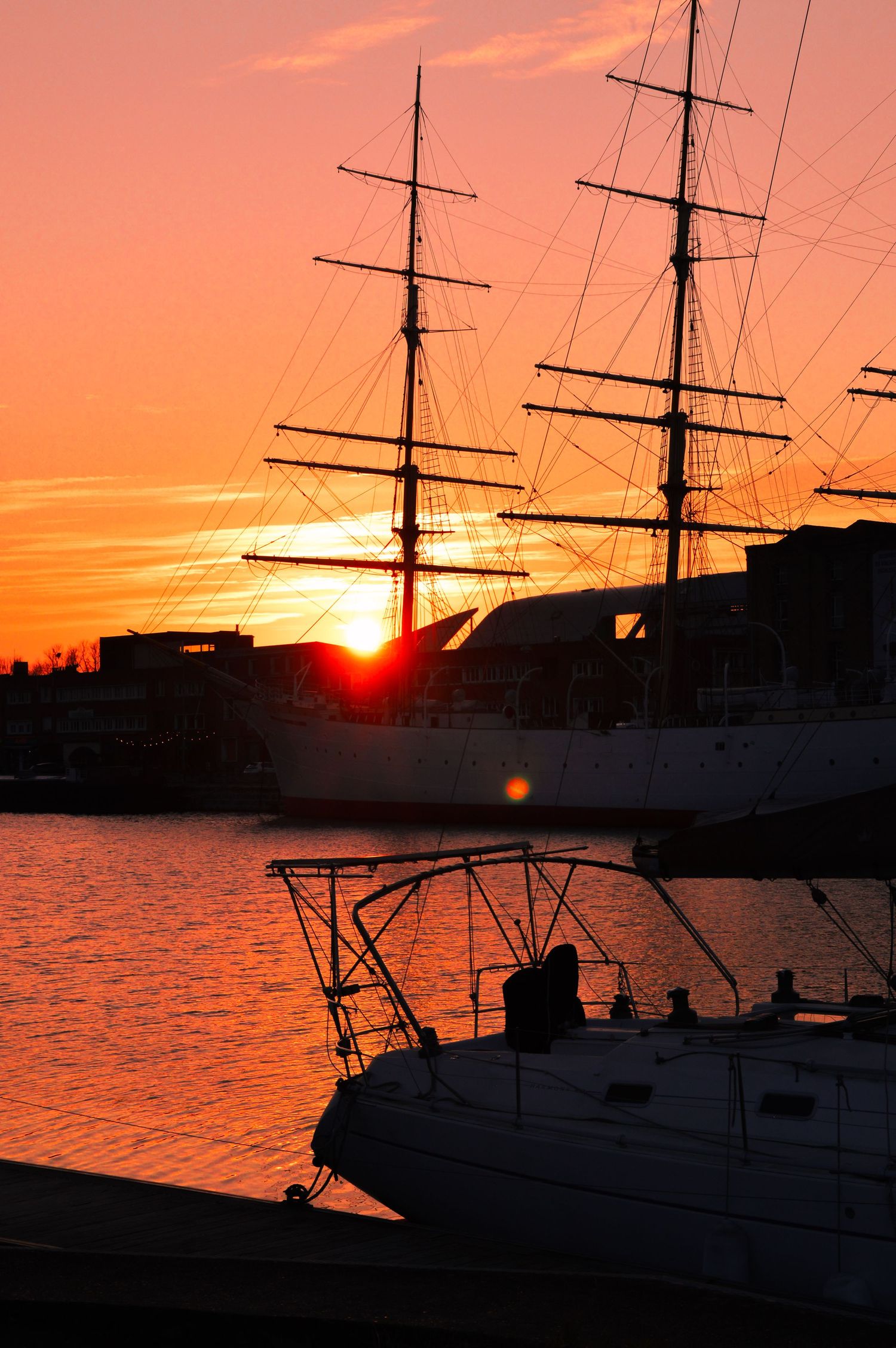 Tall ship masts and rigging outlined against brilliant orange sunset in harbor.