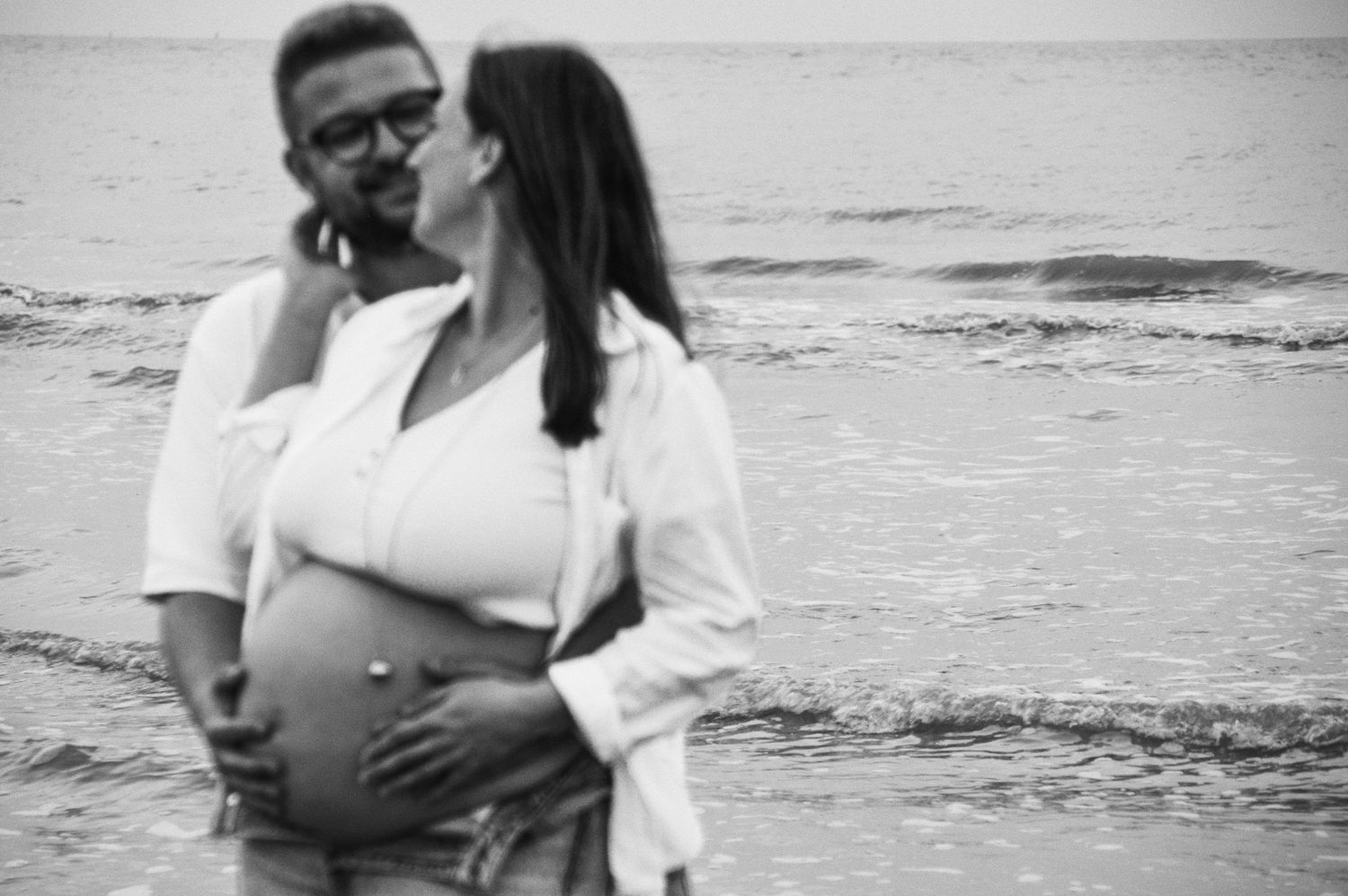 Candid beach maternity portraits in casual attire against ocean waves.