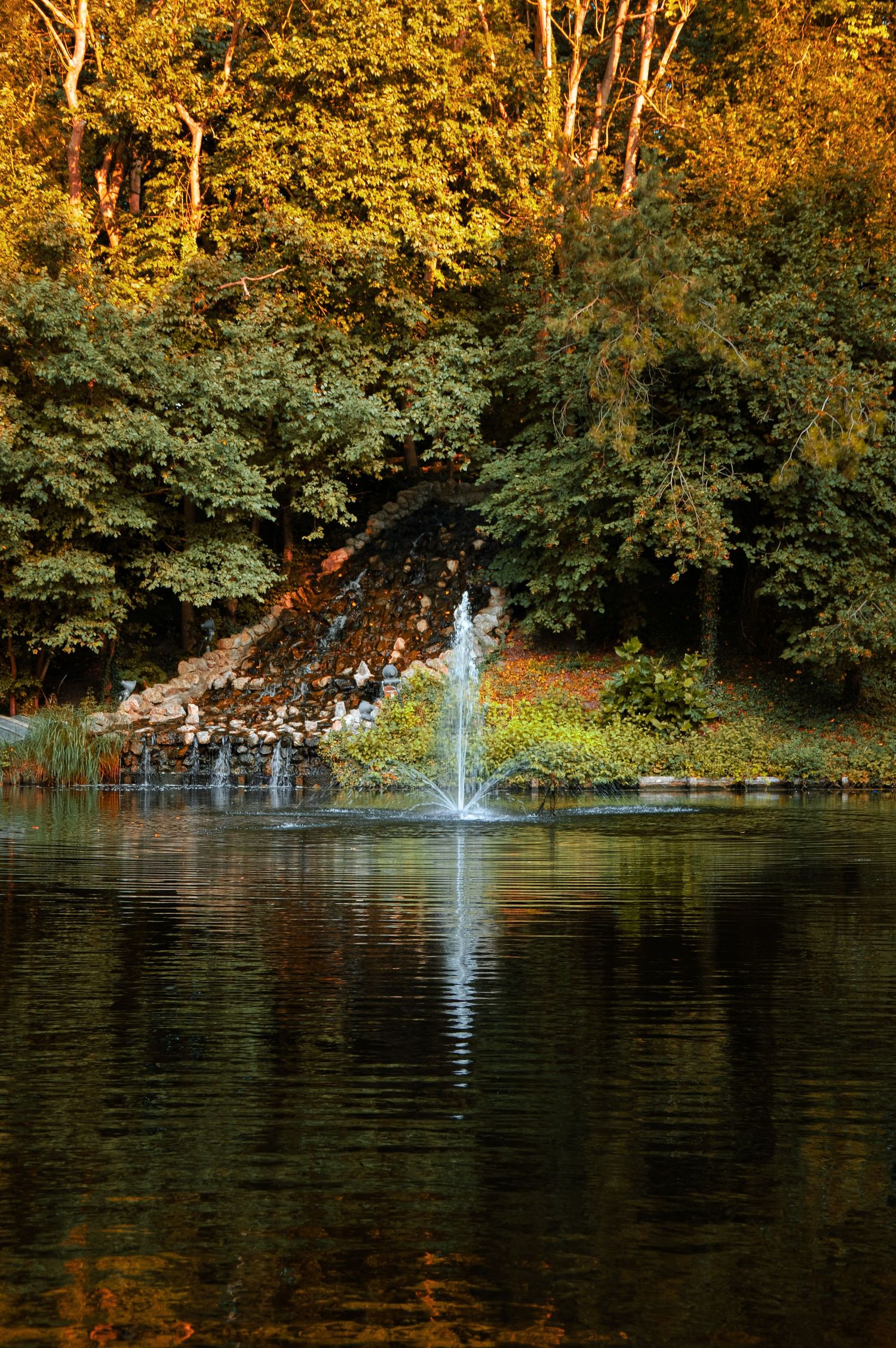 A serene pond with fountains spraying water against a backdrop of golden autumn trees.