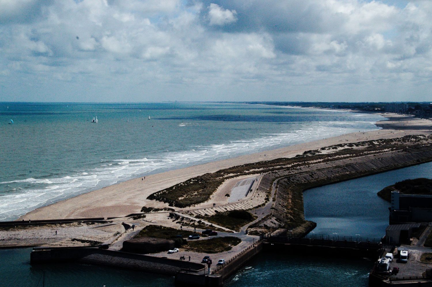 Aerial view of a coastal harbor with sandy beach, ocean waves, and cloudy sky in a scenic seaside landscape.