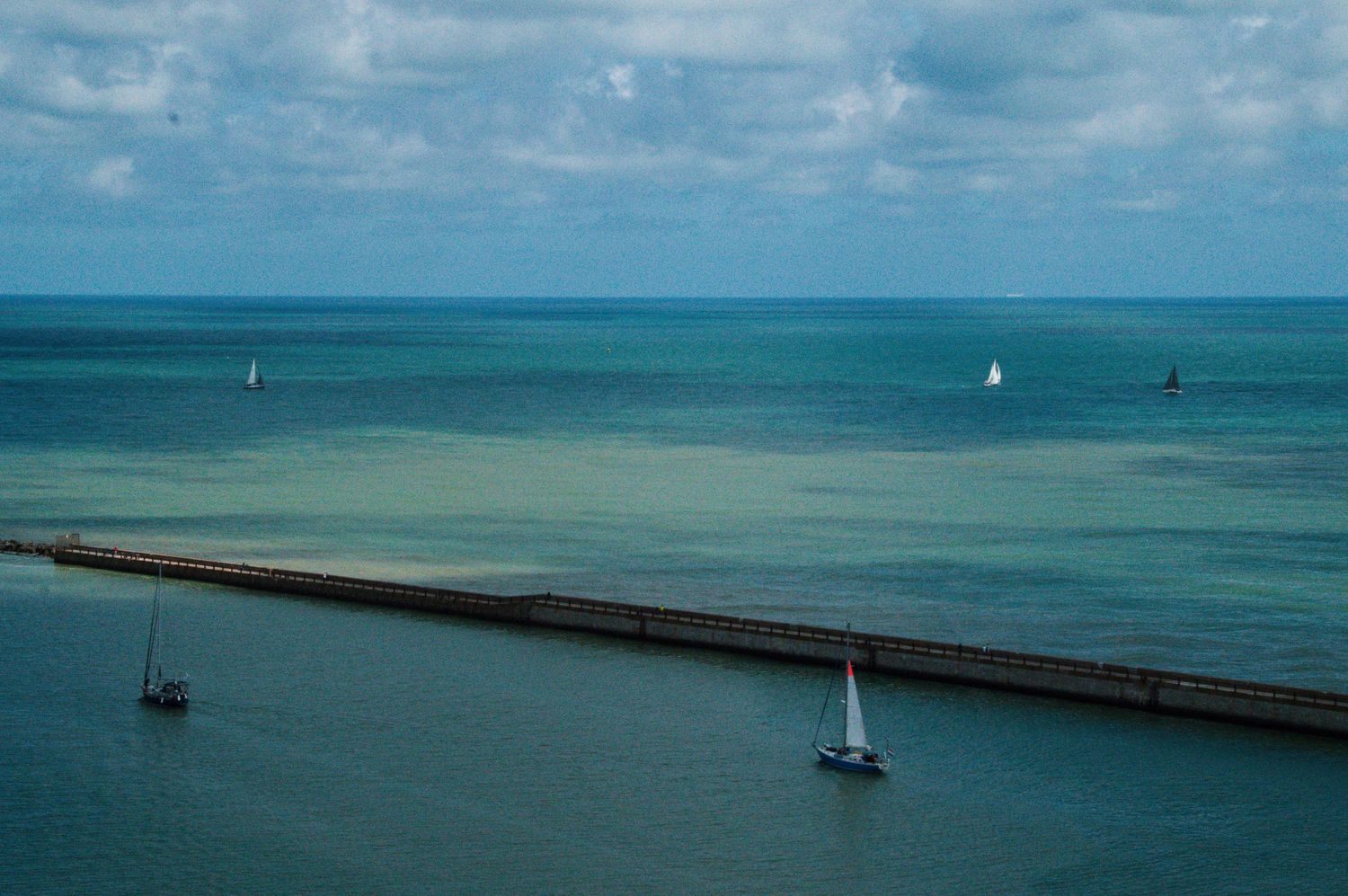Aerial view of sailboats navigating calm turquoise waters near a long pier under a cloudy sky.