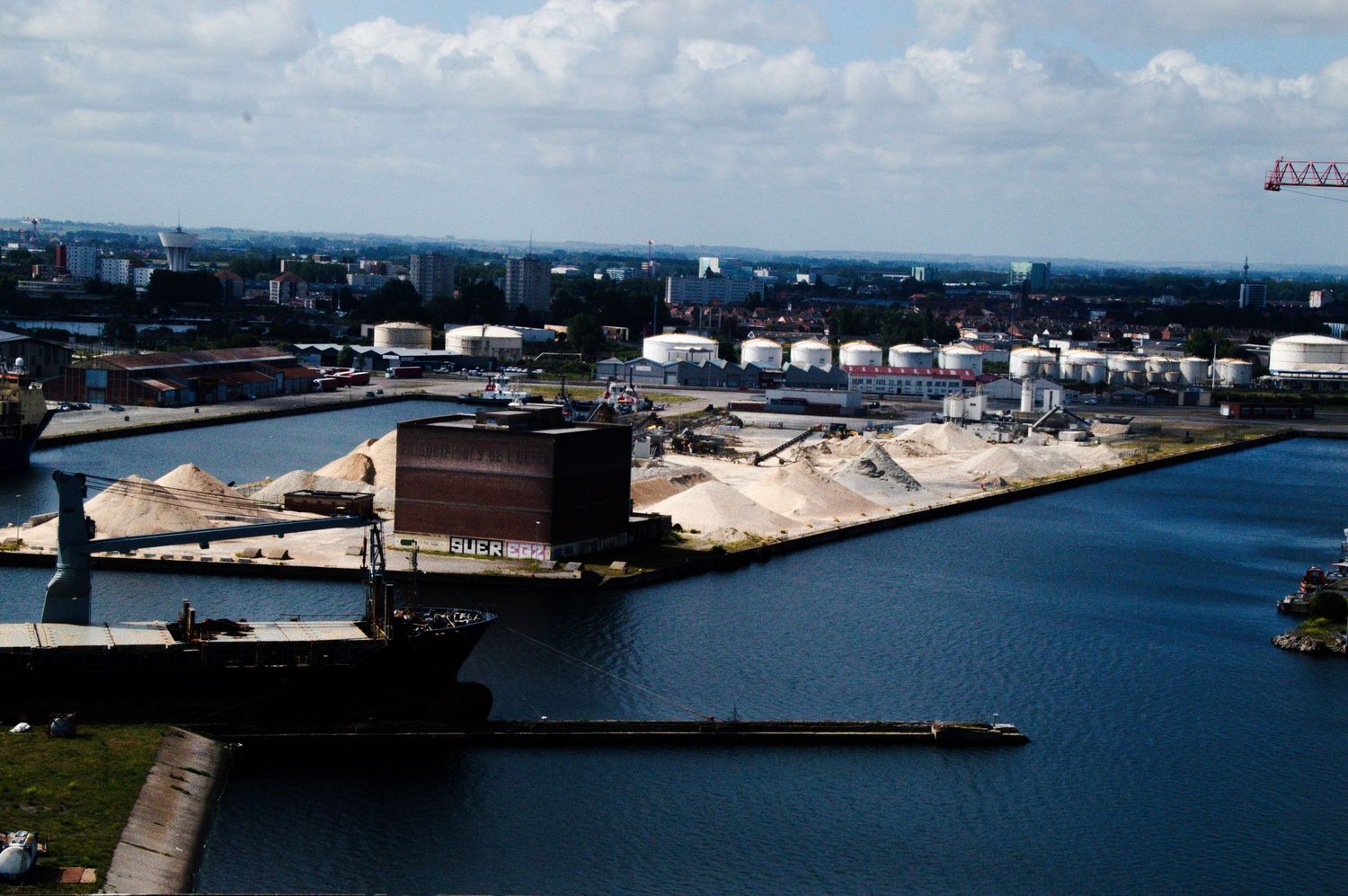 Aerial view of an industrial harbor with storage tanks, bulk material piles, and a calm blue waterway under partly cloudy skies.