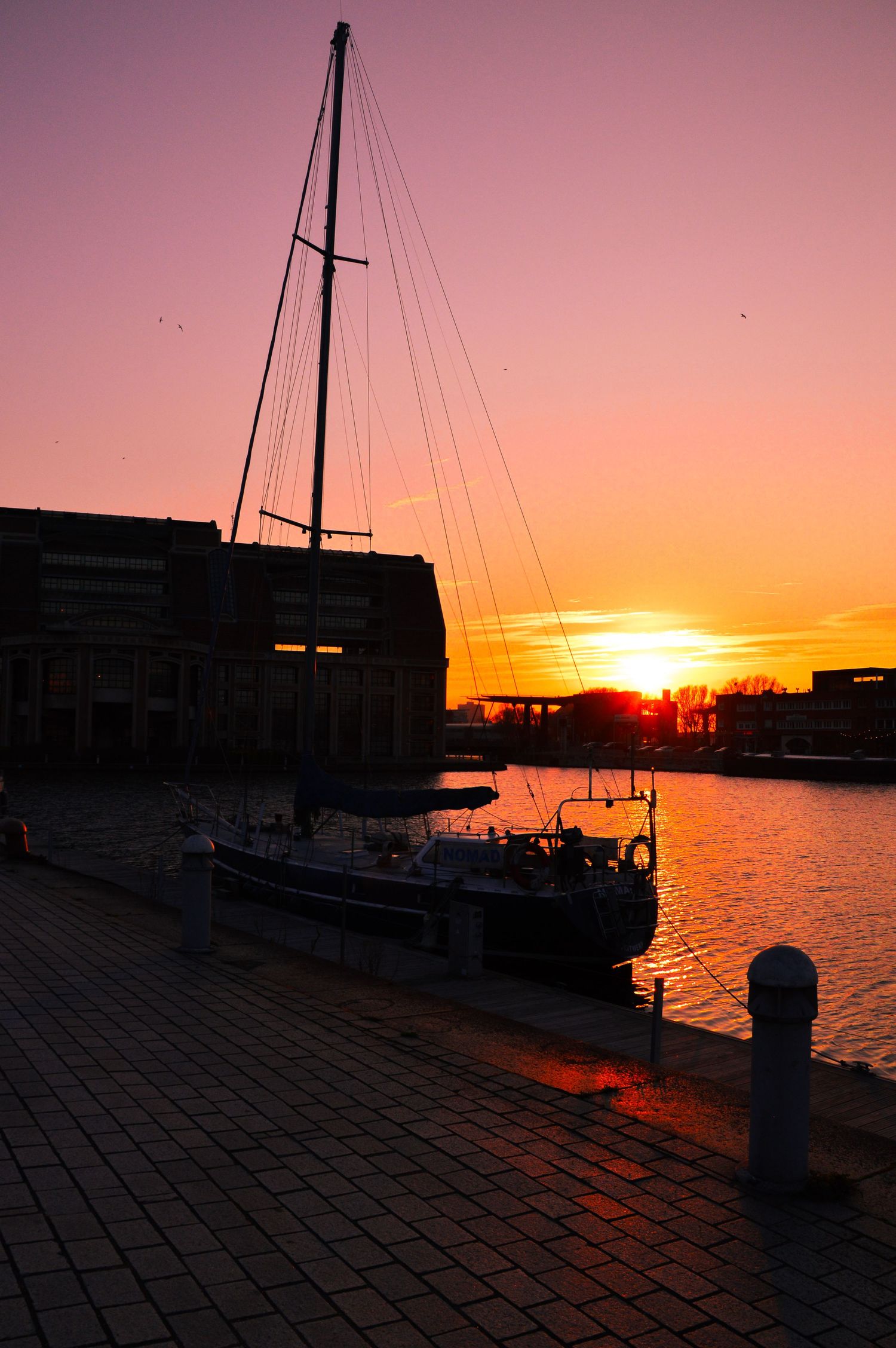 Waterfront dock with moored sailboat silhouetted against brilliant orange sunset.