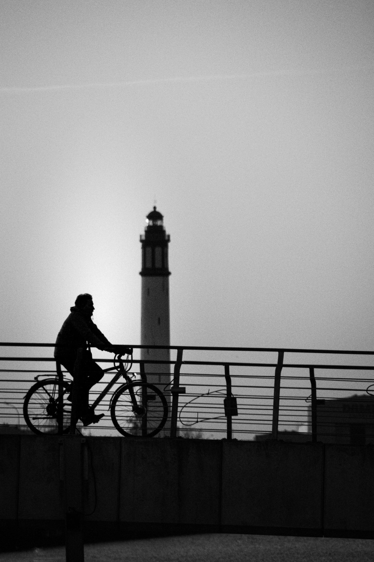 A silhouetted cyclist rides along a railing with a lighthouse in the background at dusk.