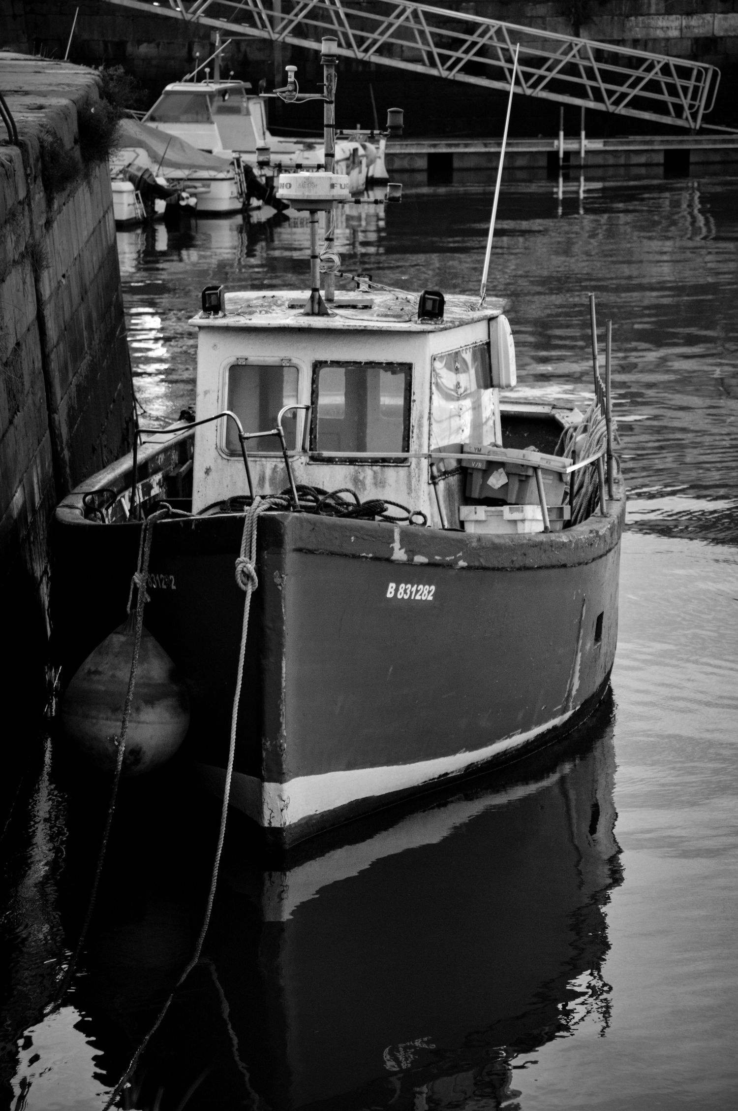Black and white image of fishing boats moored at dock with reflections in water.