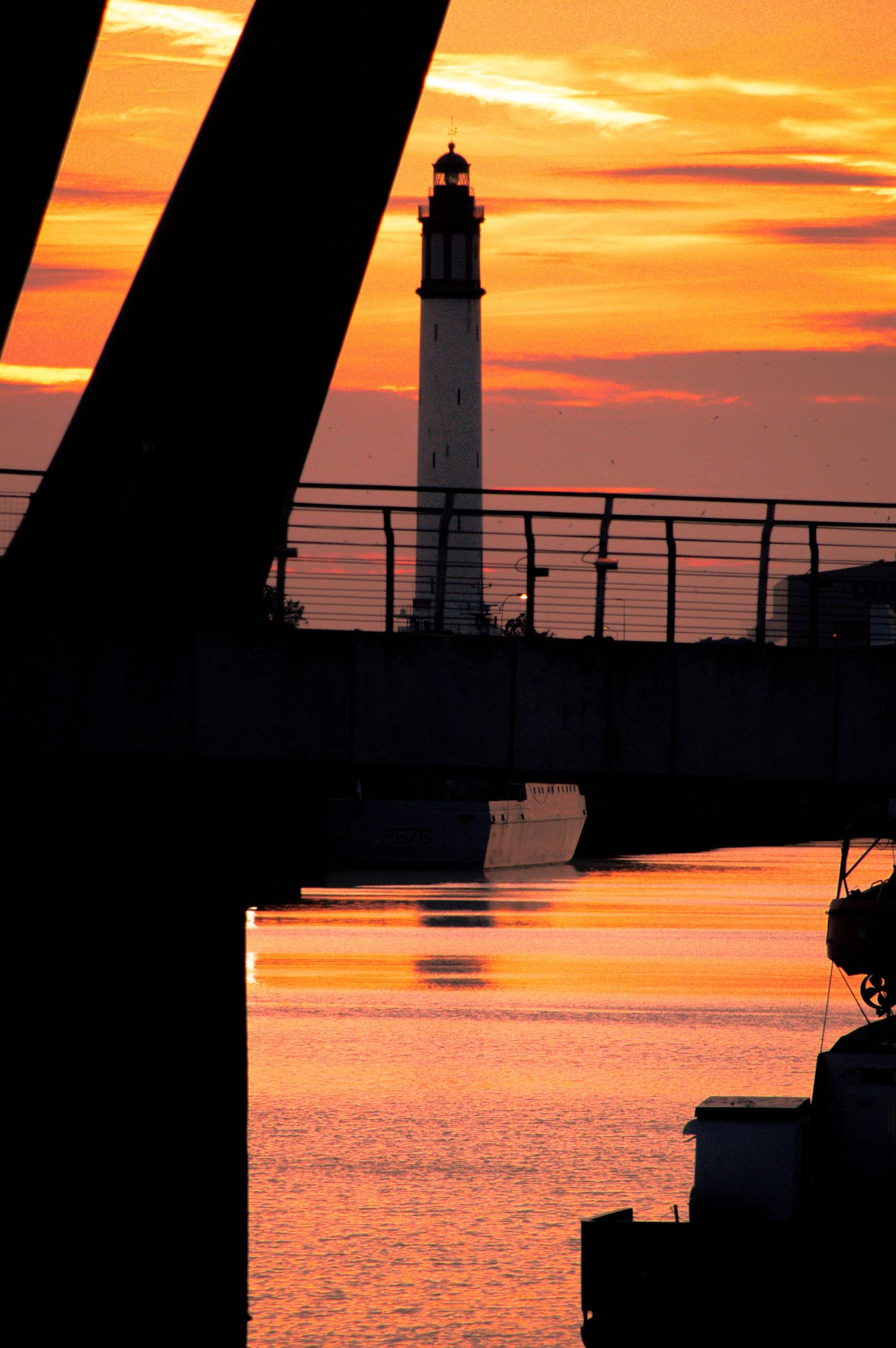 Silhouette of a lighthouse at sunset with vibrant orange sky reflected on calm harbor waters framed by bridge structure.