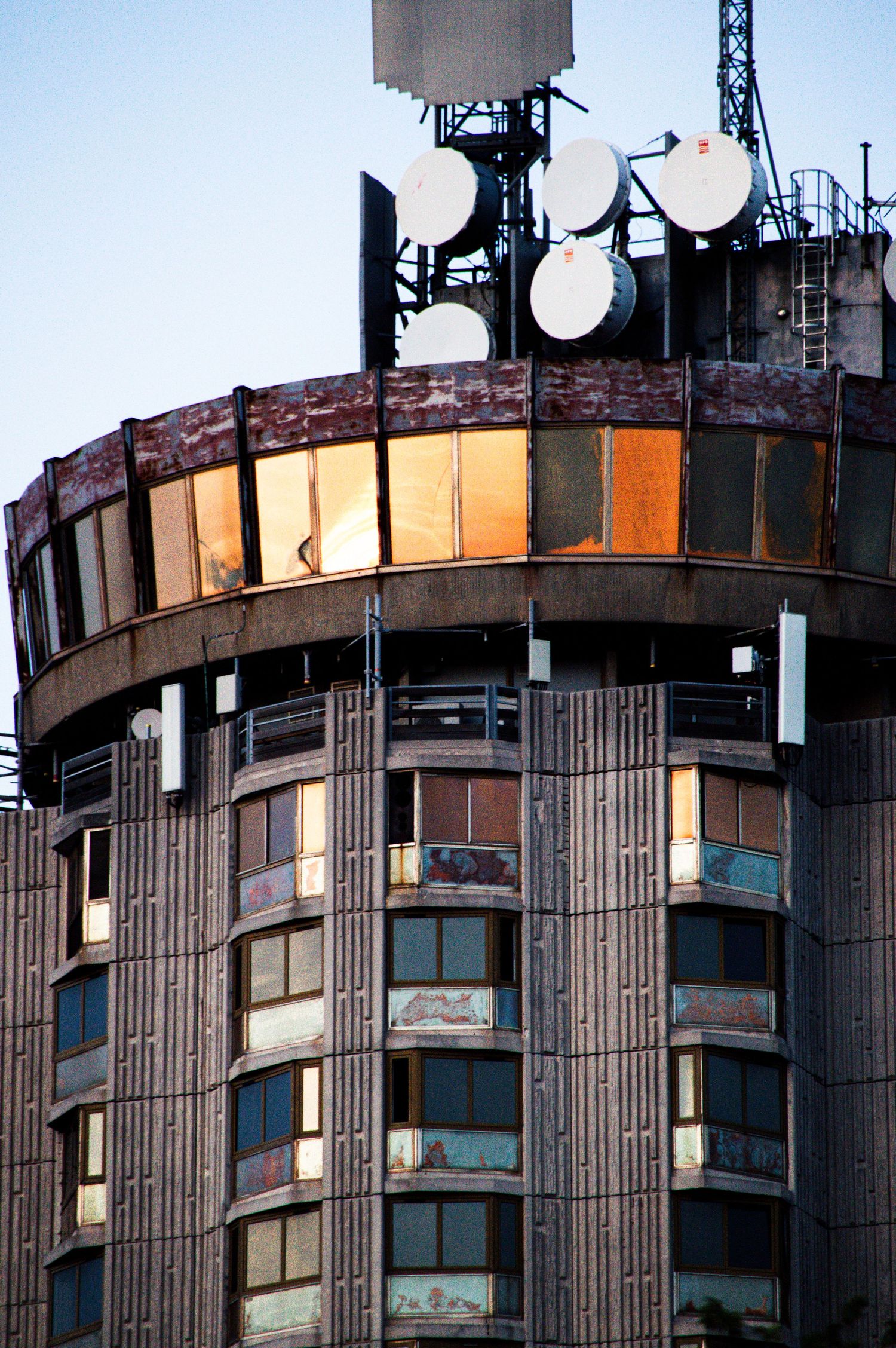Brutalist cylindrical skyscraper with satellite dishes on rooftop, reflective windows glowing at sunset.